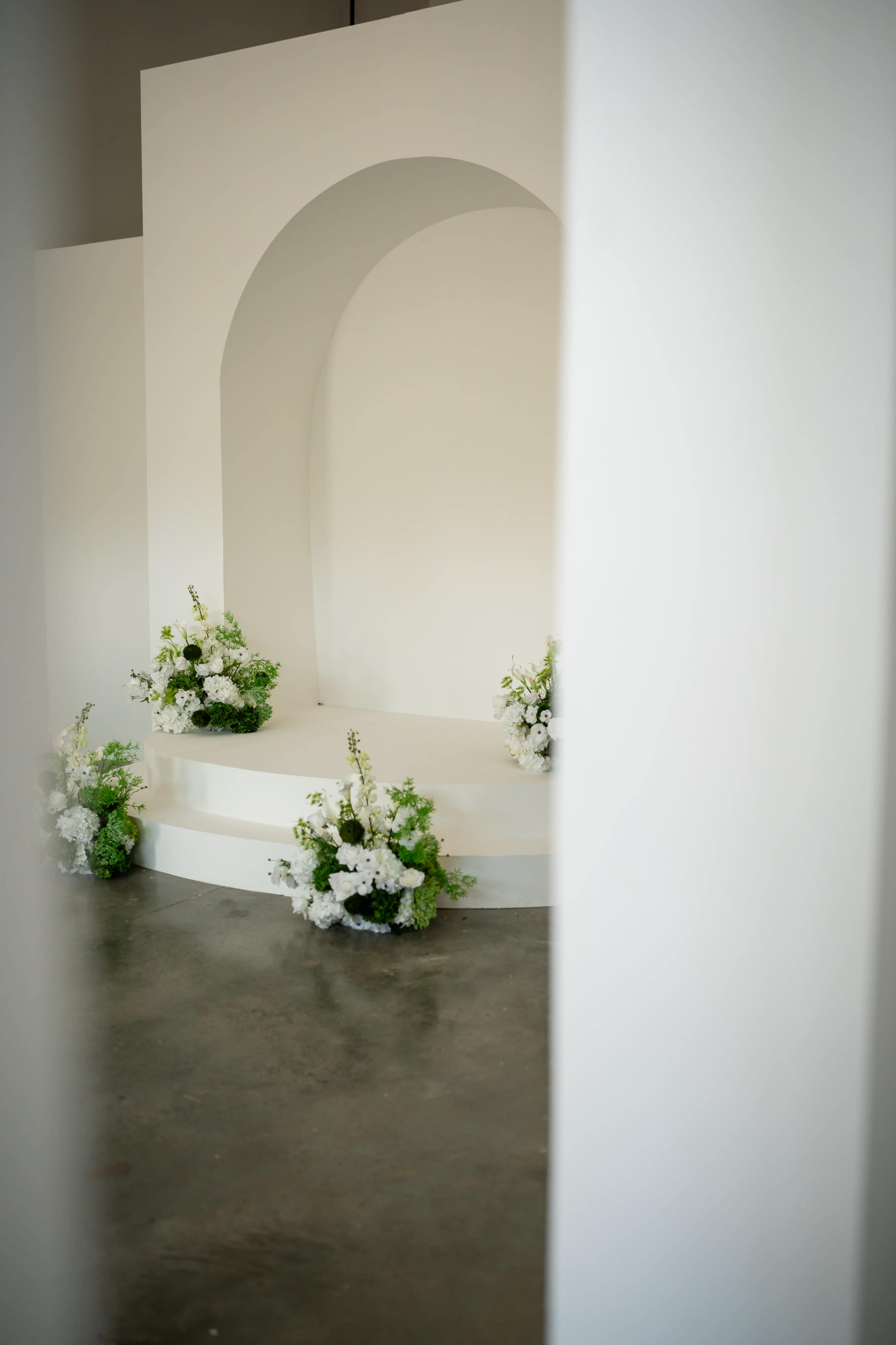 The image shows a minimalist stage with a curved arch and several flower arrangements on either side.
