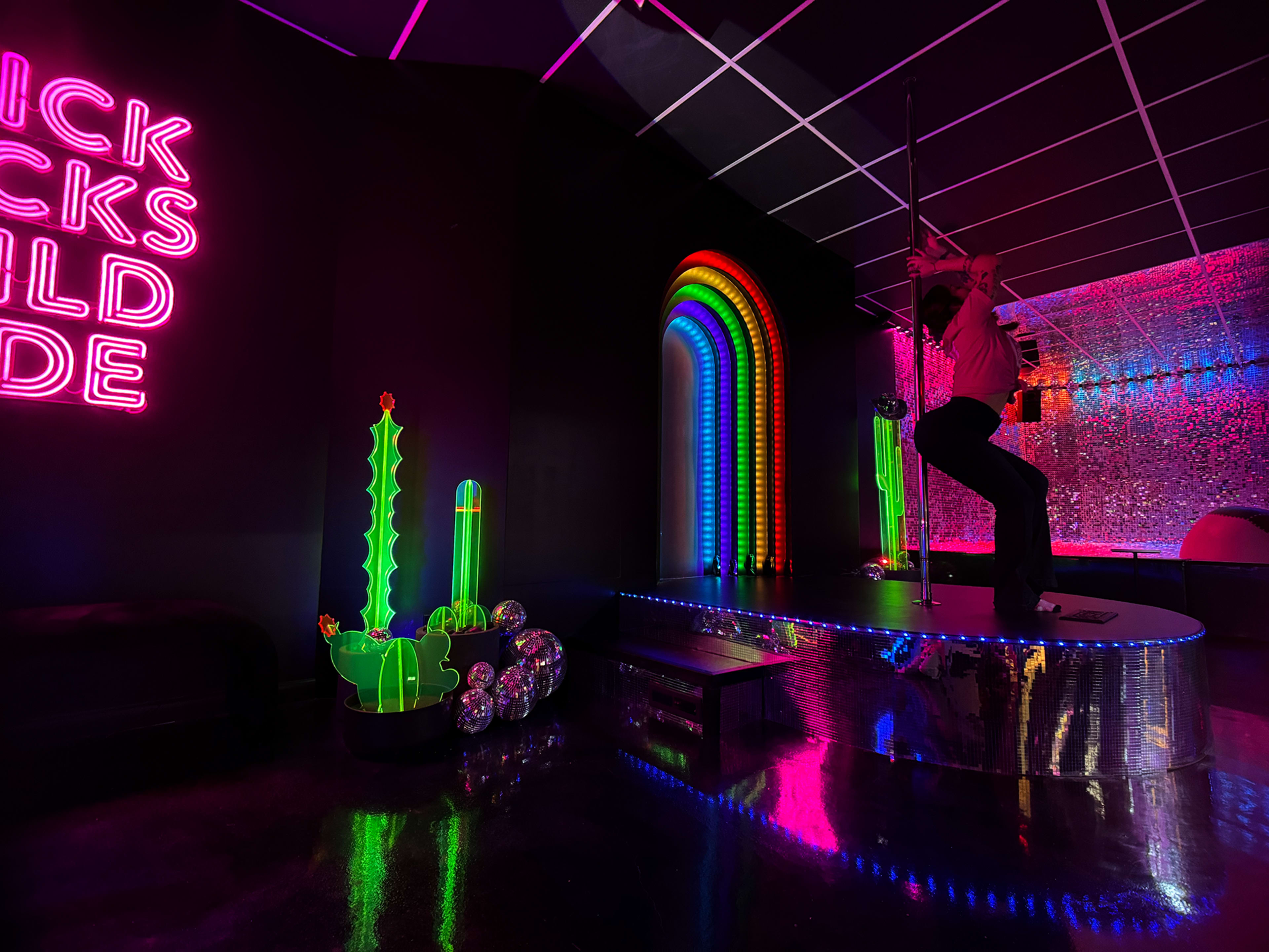 The scene features a vibrant, neon-lit room with colorful decorations, including a rainbow arch, a cactus sculpture, and a pole dancer on a stage.