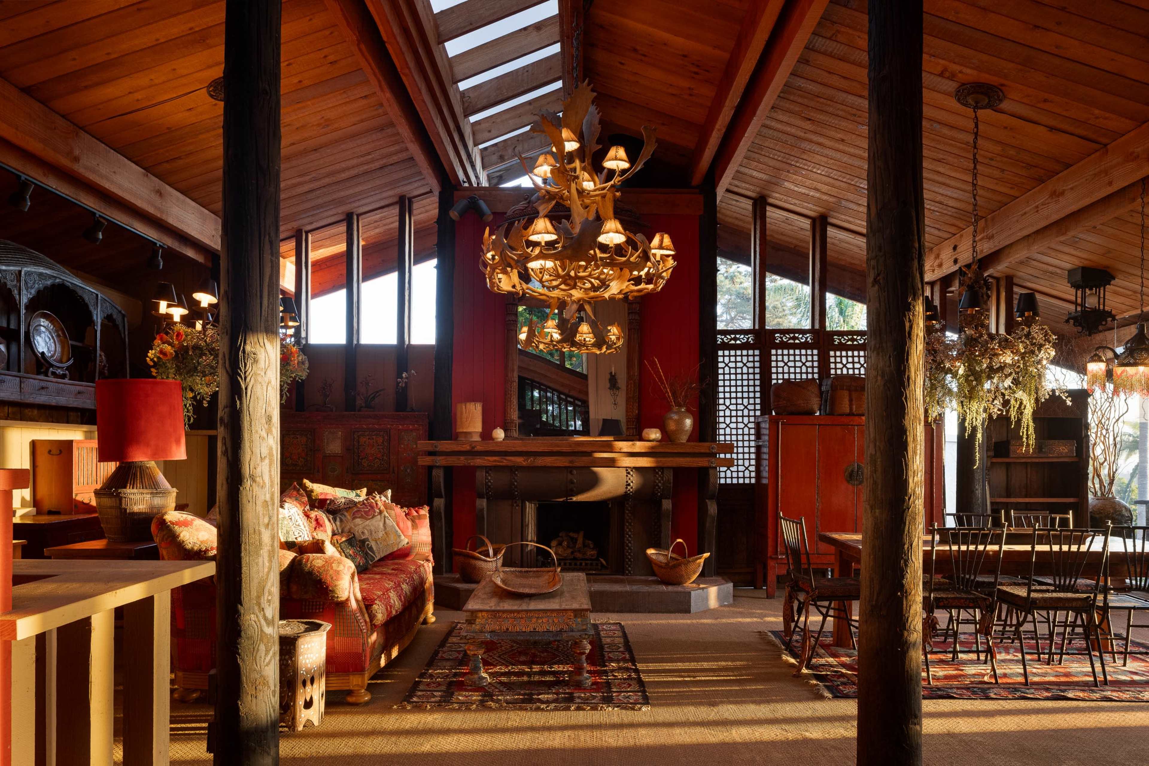 The image shows a spacious wooden interior of a rustic living room and dining area, featuring a large chandelier, a fireplace, and various decorative furnishings.