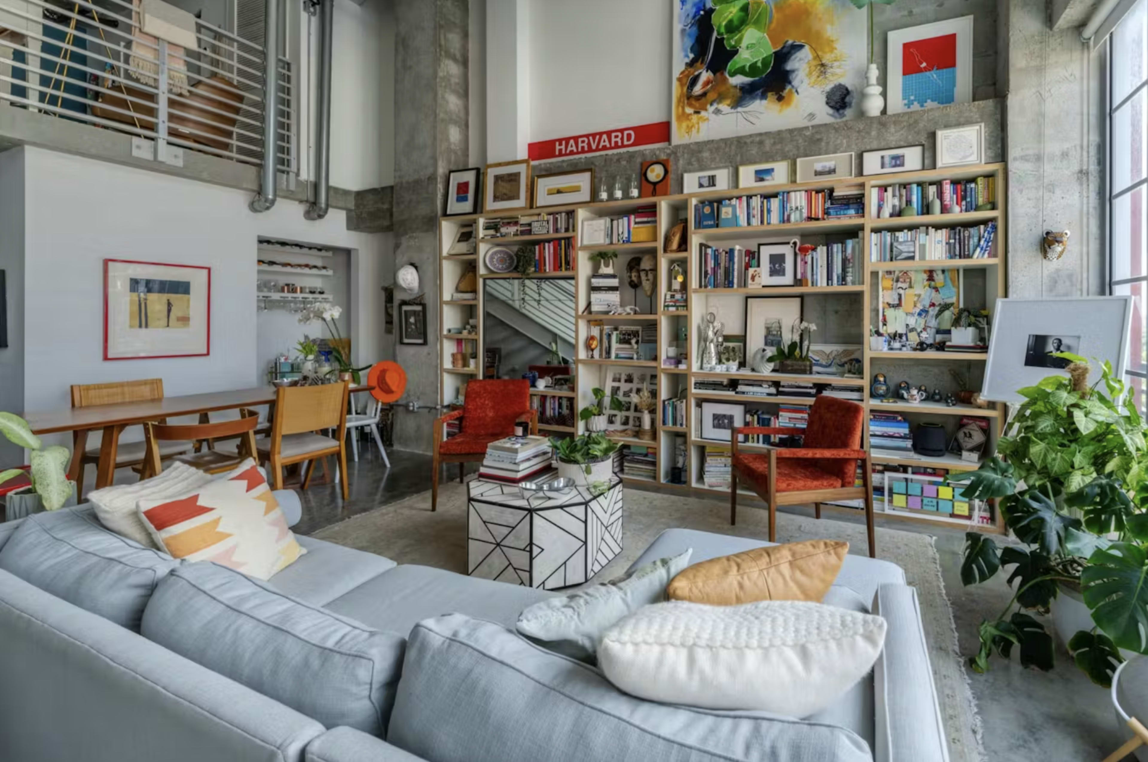A modern living room with a light gray sofa, a patterned coffee table, a bookshelf filled with books and decorative items, and two red chairs, all set in a loft-style space with large windows.