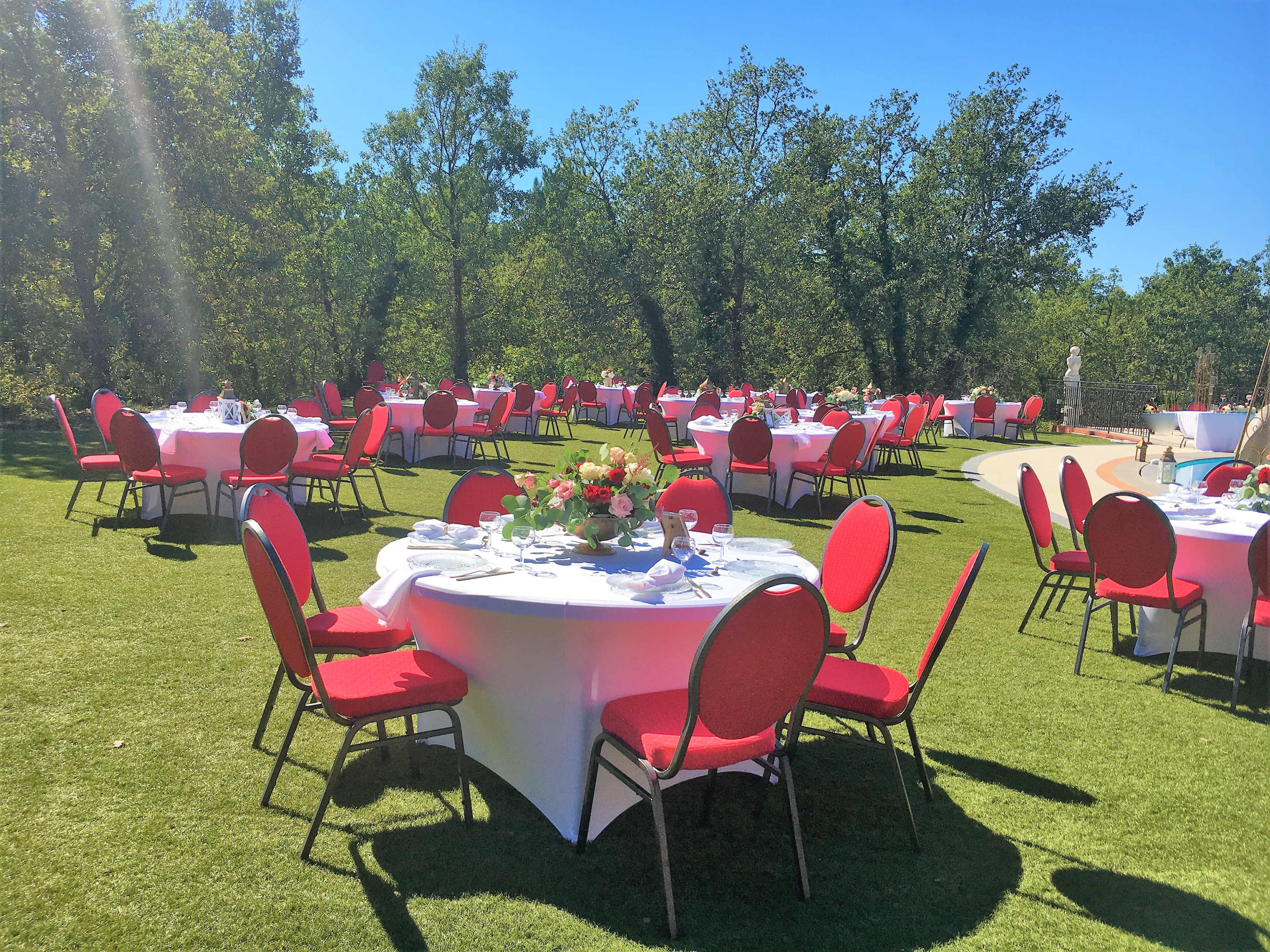 The image shows a landscaped outdoor area with several round tables set for dining, featuring white tablecloths and vibrant red chairs under a clear blue sky.