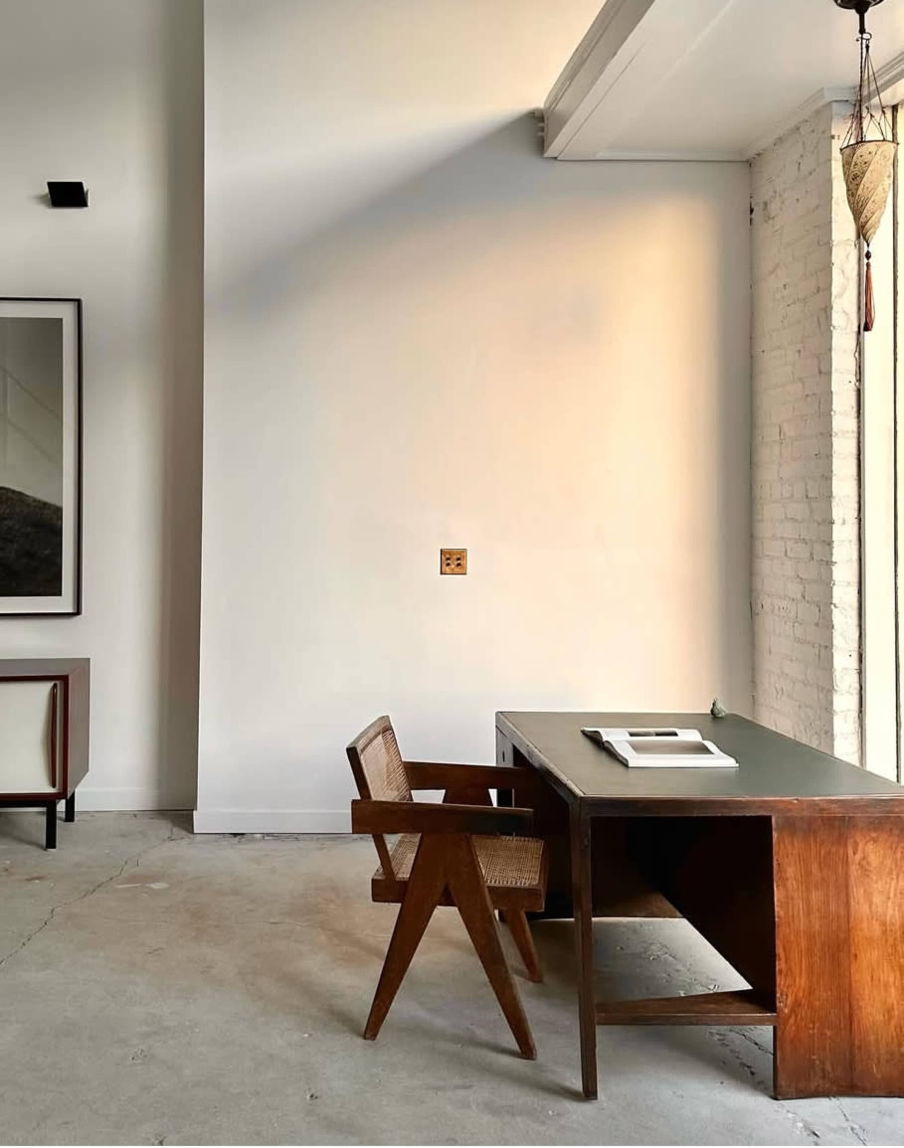 The image shows a minimalist office space featuring a wooden desk and a chair against a blank wall, with a large window allowing light to enter.