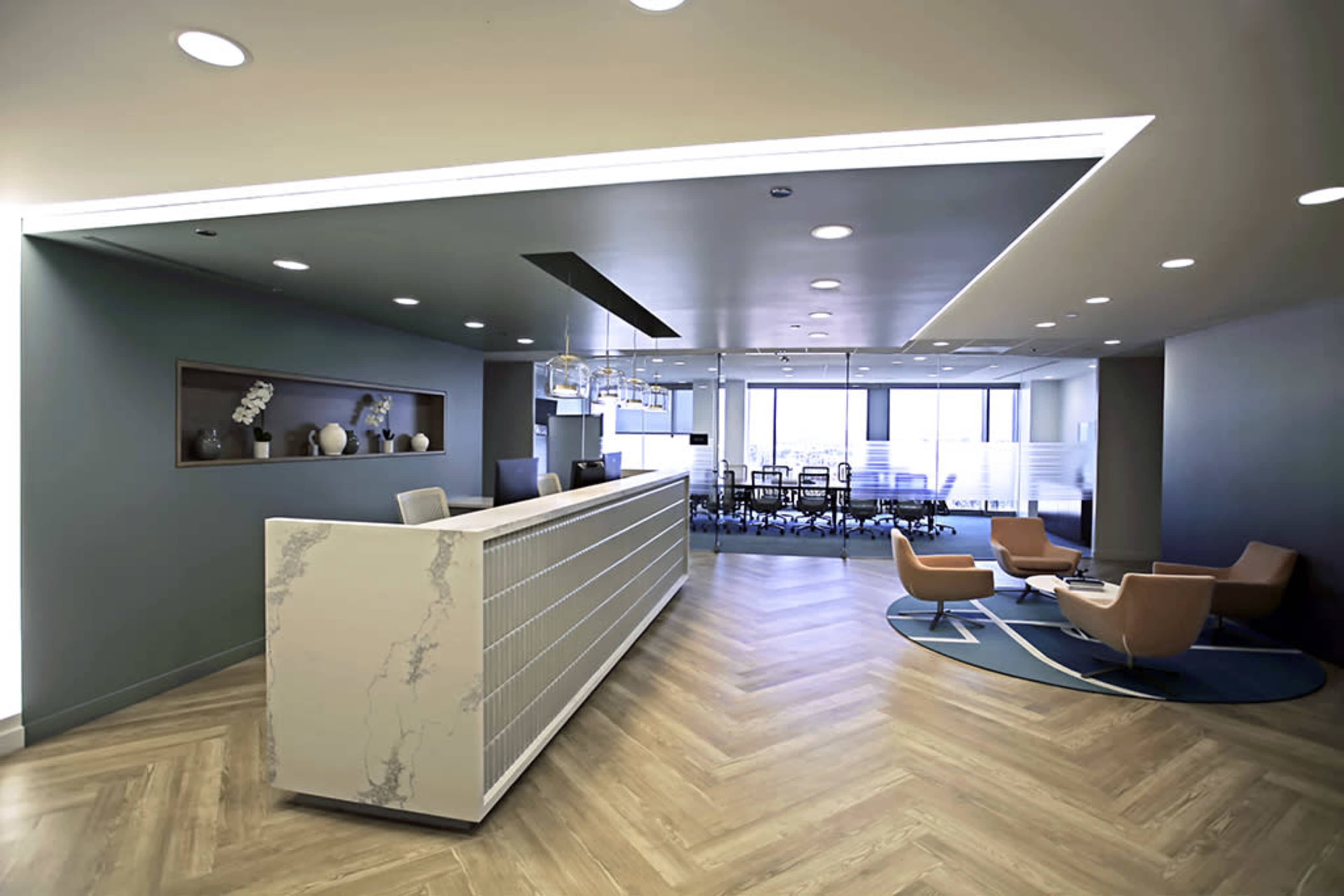 A modern office reception area features a marble front desk, seating arrangements with orange chairs, and a conference room visible in the background.