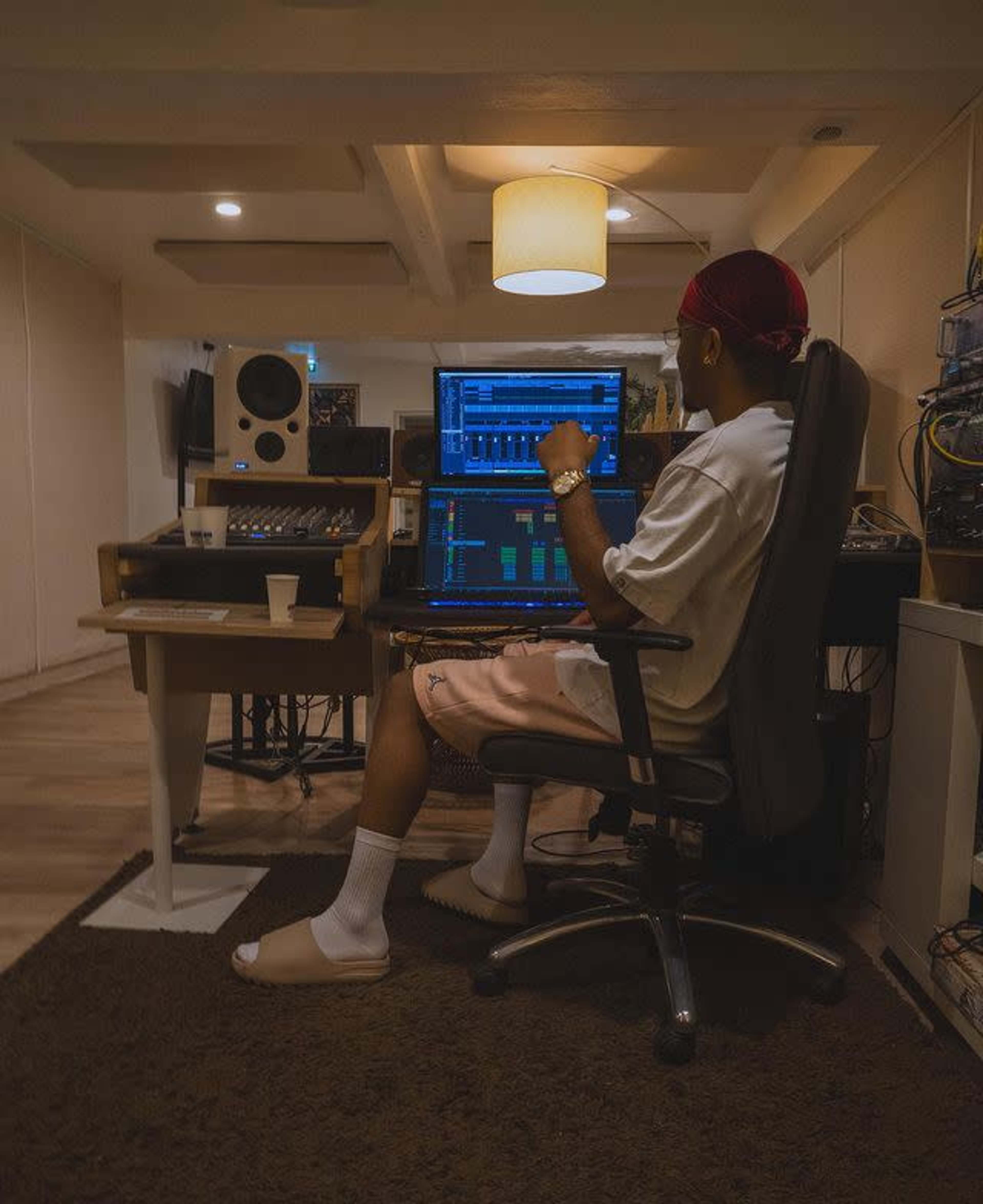 A person sits in a chair in a music studio, working at a computer with sound equipment and monitors.