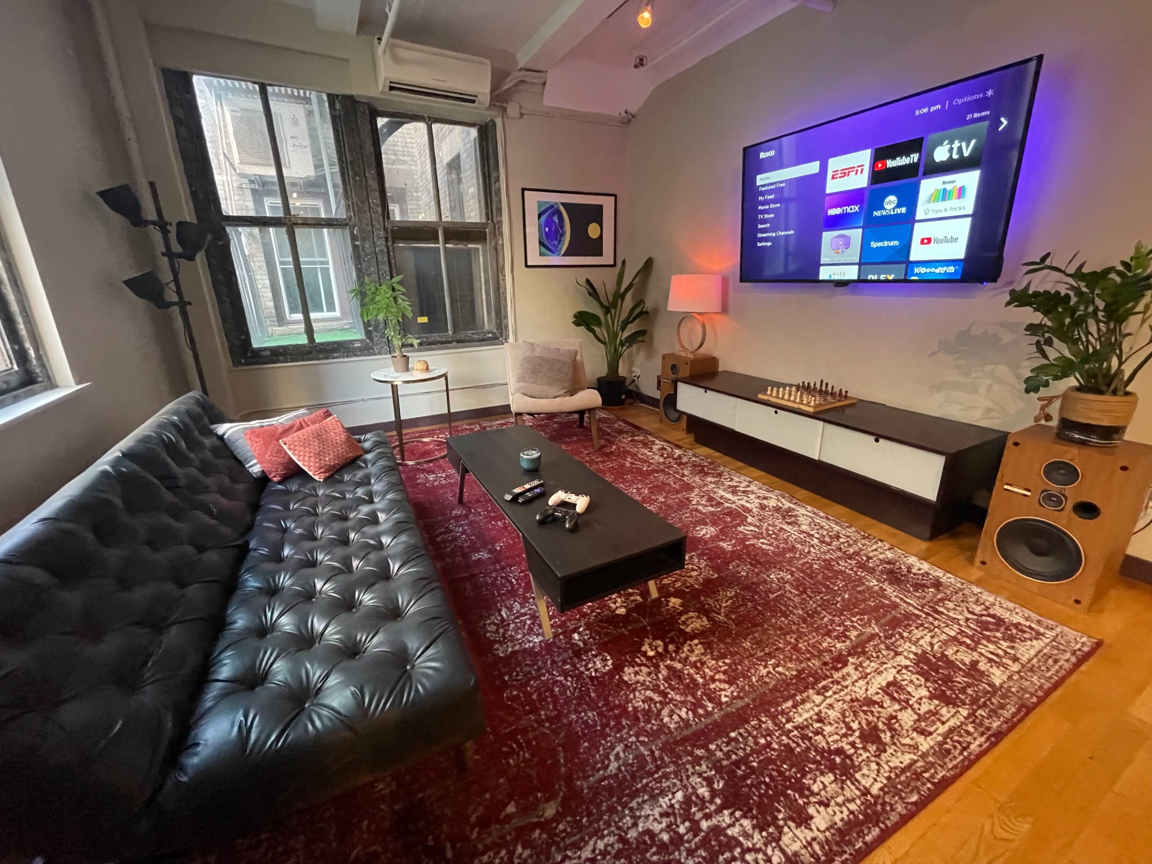 A cozy living room with a tufted black sofa, a coffee table, and a large TV mounted on the wall, surrounded by natural light from two windows.