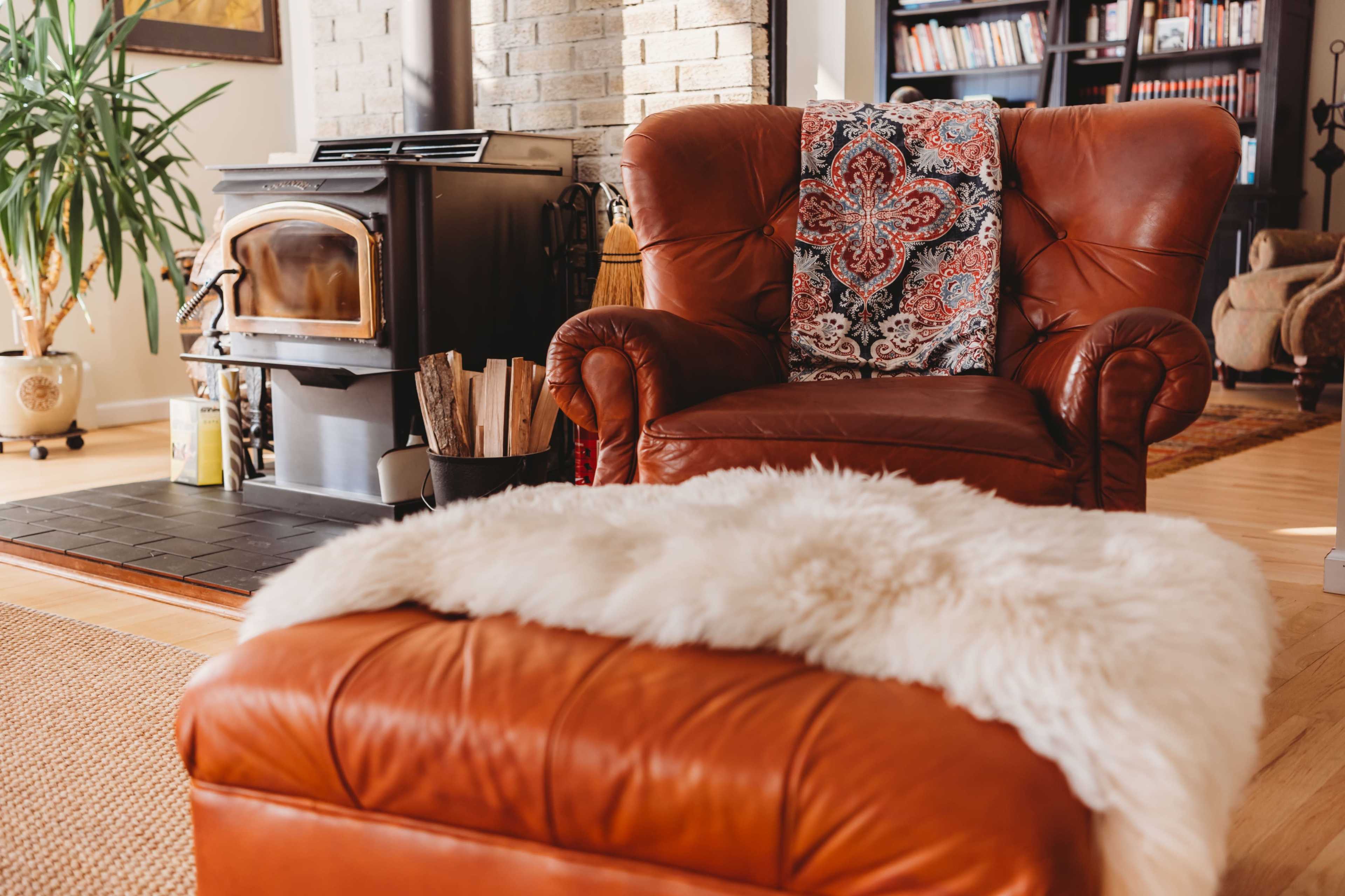 The image shows a cozy living room with a brown leather armchair, a wood stove, and a fur-covered ottoman.