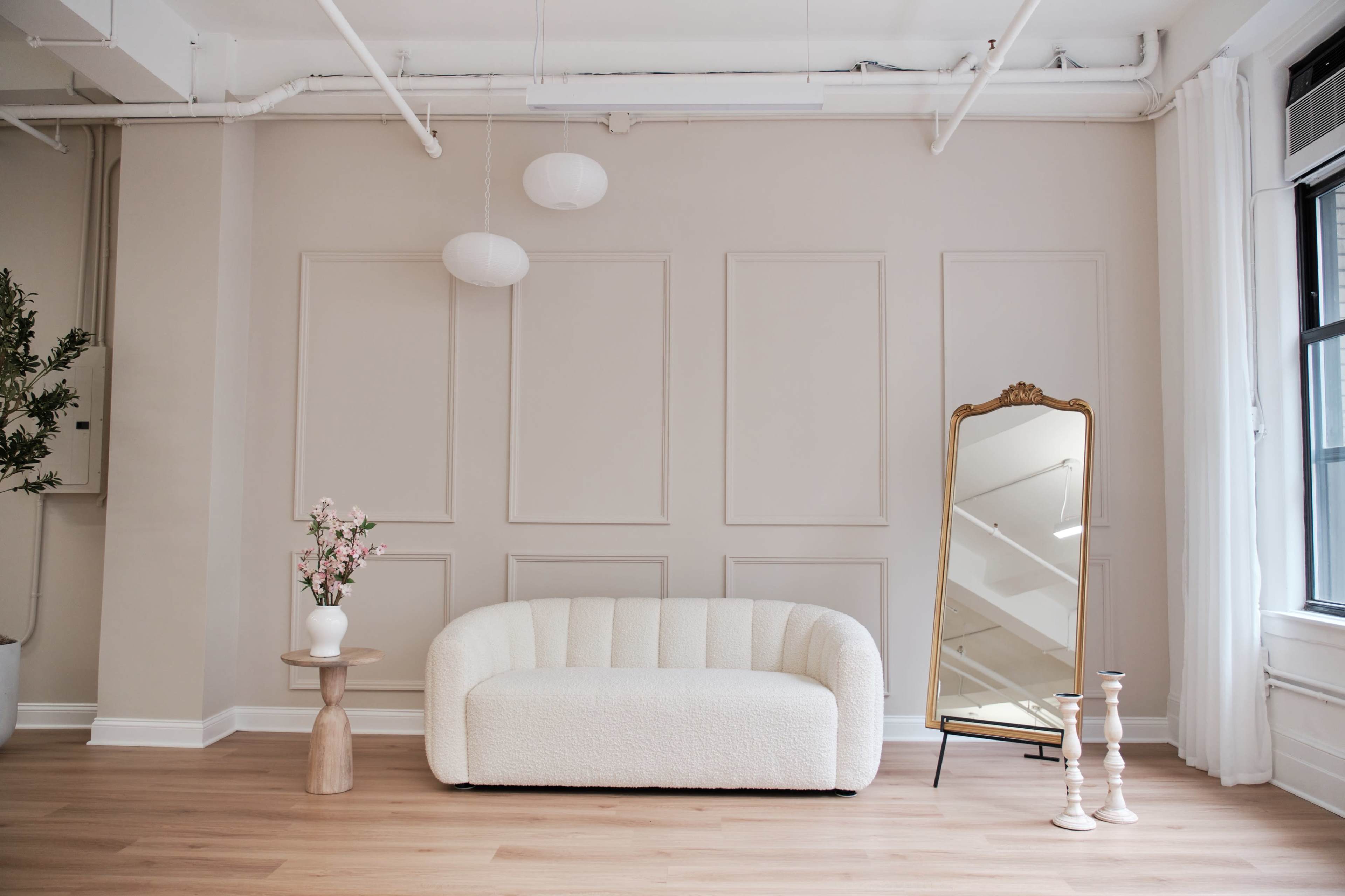 The room features a white sofa, a mirror standing on a black frame, and a vase with flowers on a wooden table, all set against a beige wall with paneling.
