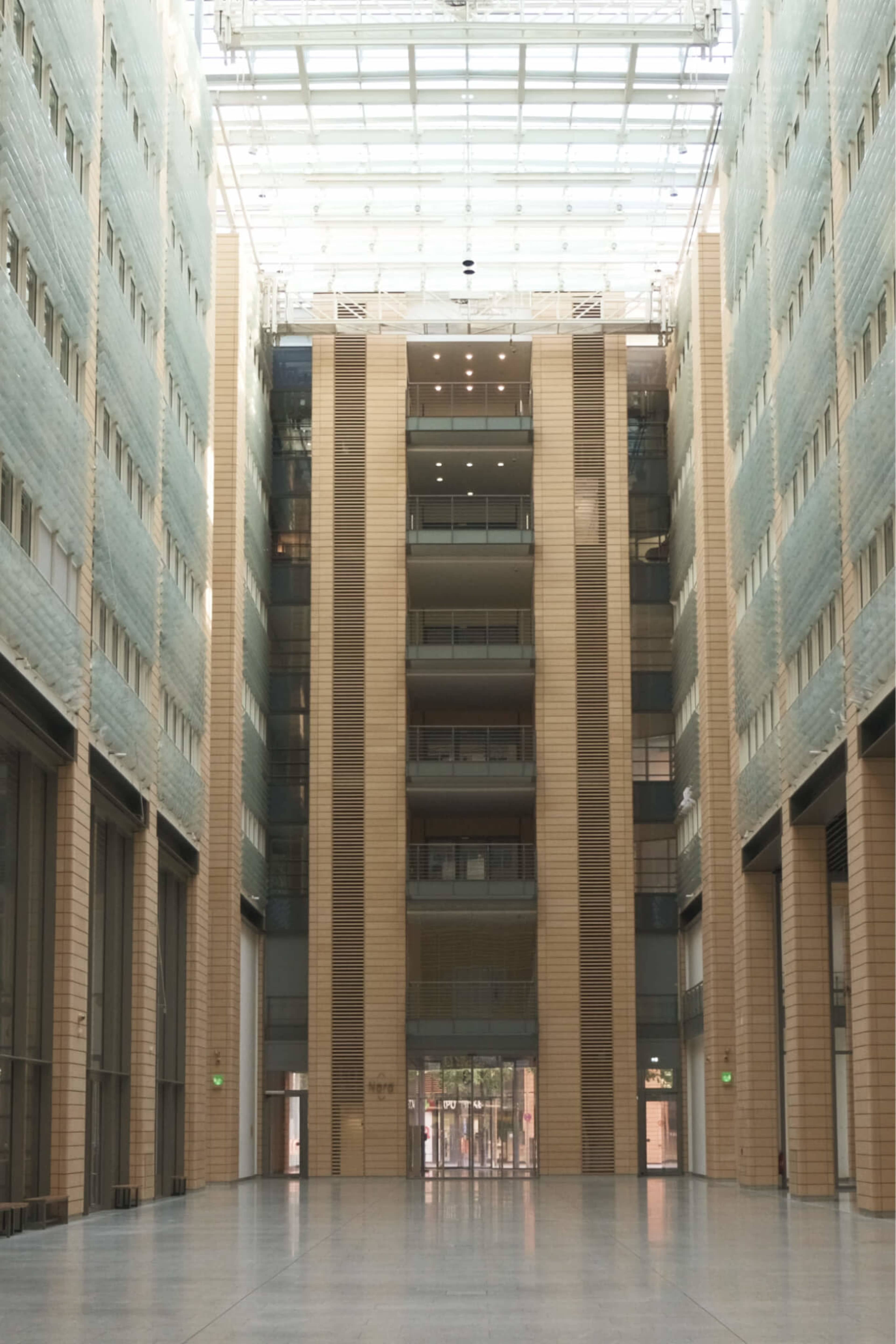 The image shows a large, modern atrium with tall glass walls and a central staircase flanked by beige columns.