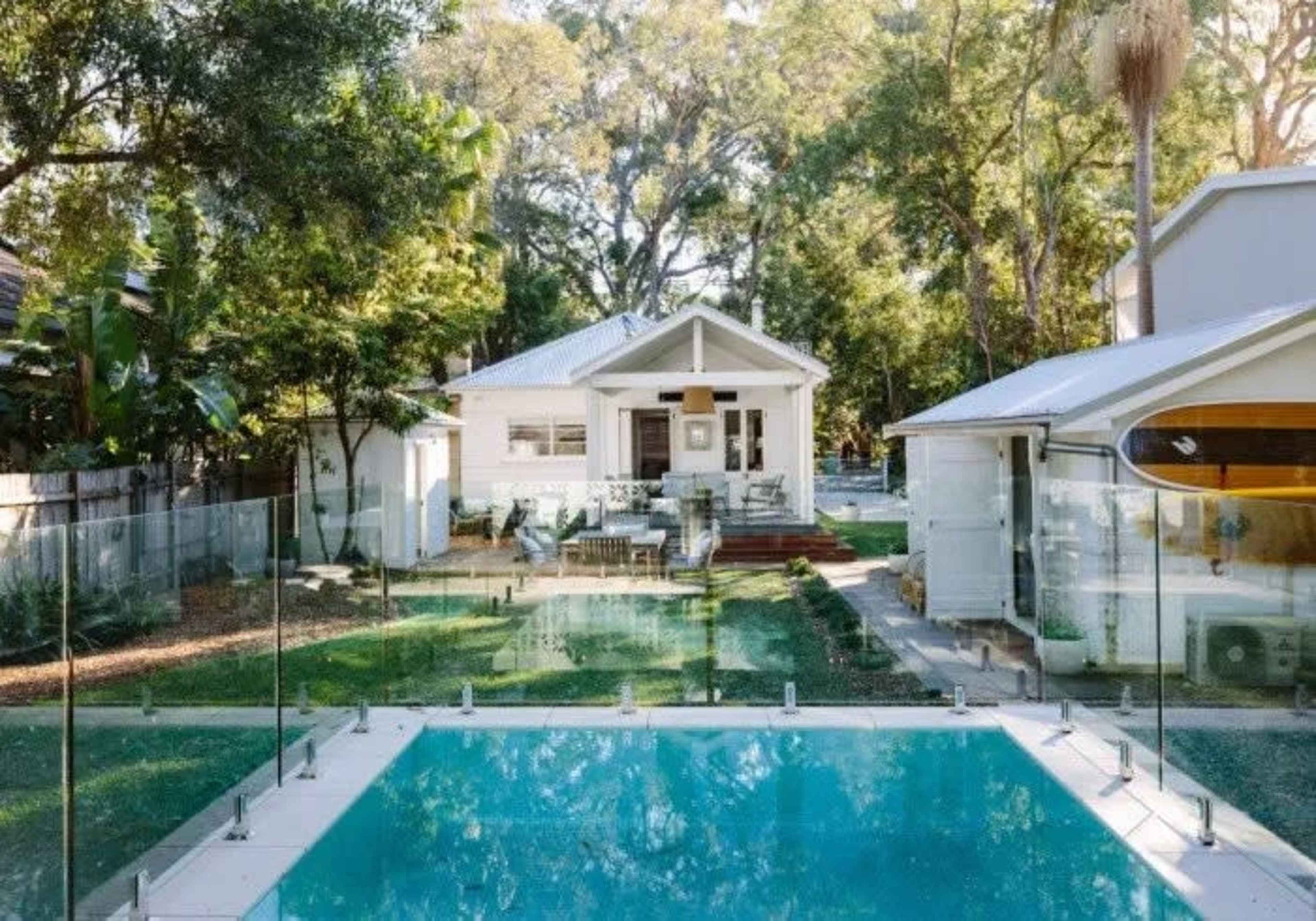 A clear swimming pool is situated in front of a modern house surrounded by greenery and a glass fence.