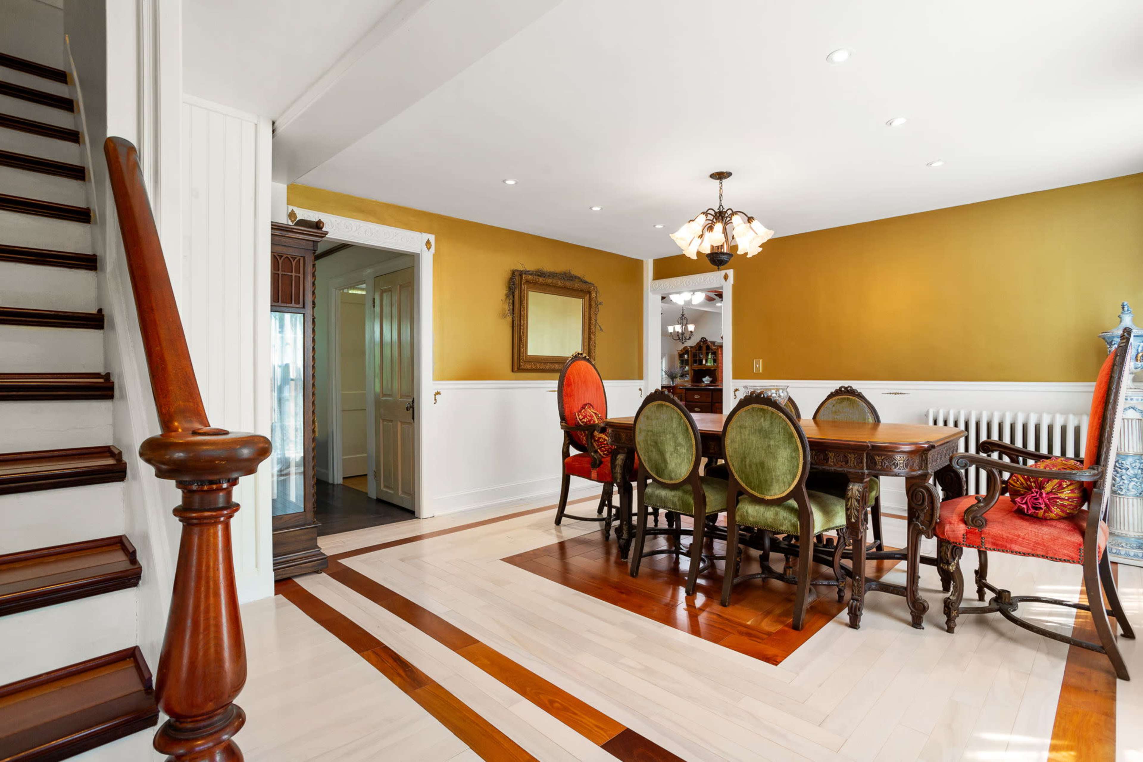 A dining area with a wooden table surrounded by chairs, a staircase on the left, and a wall painted in a warm gold color.