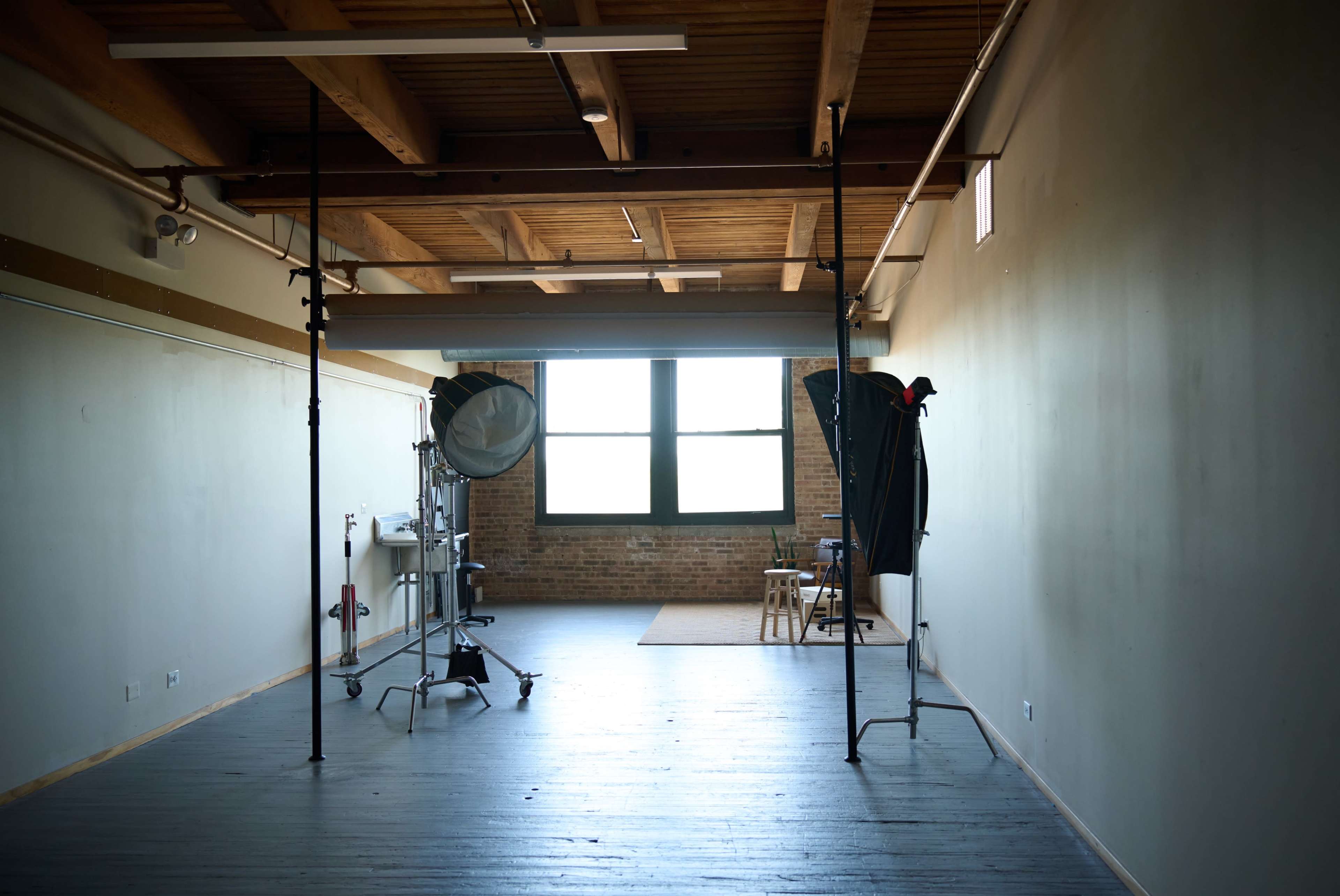 The image shows a spacious, empty photo studio with wooden beams and two lighting setups on either side.