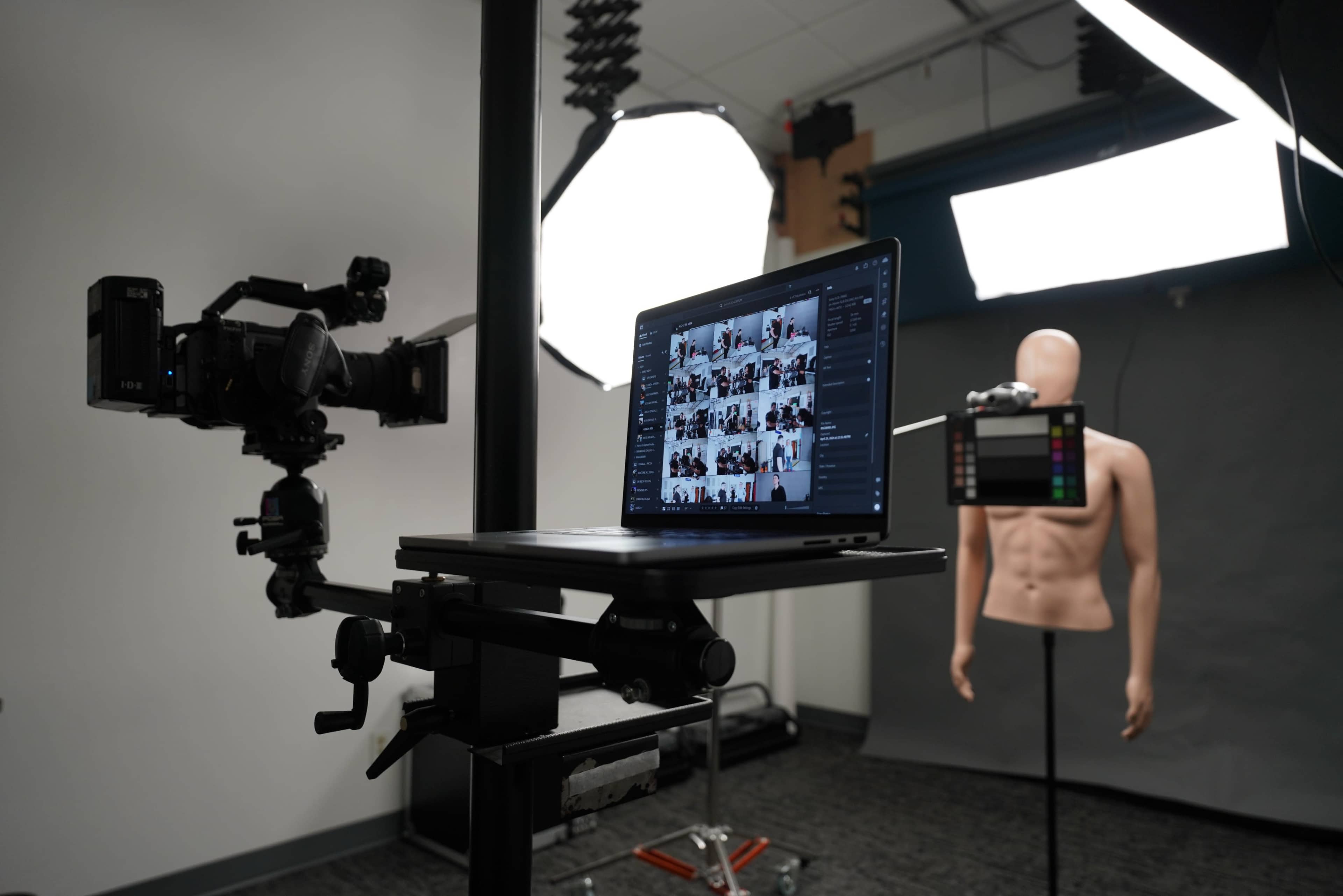 A camera and a laptop are positioned on a stand in a photography studio with a mannequin and lighting equipment set up in the background.
