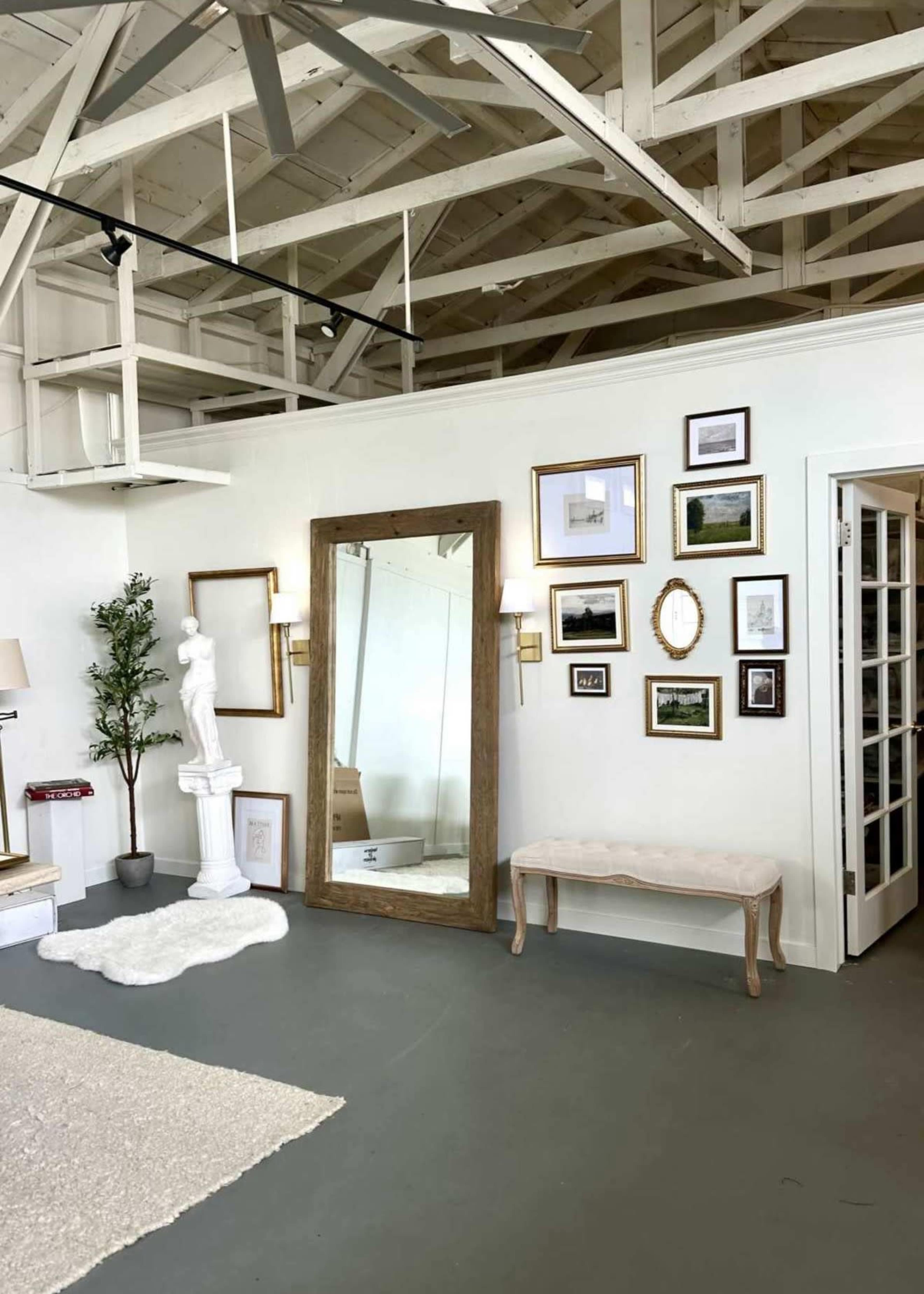 The image shows an interior space with a large mirror, framed photographs on the wall, a statue, a bench, and a light-colored rug on the floor.