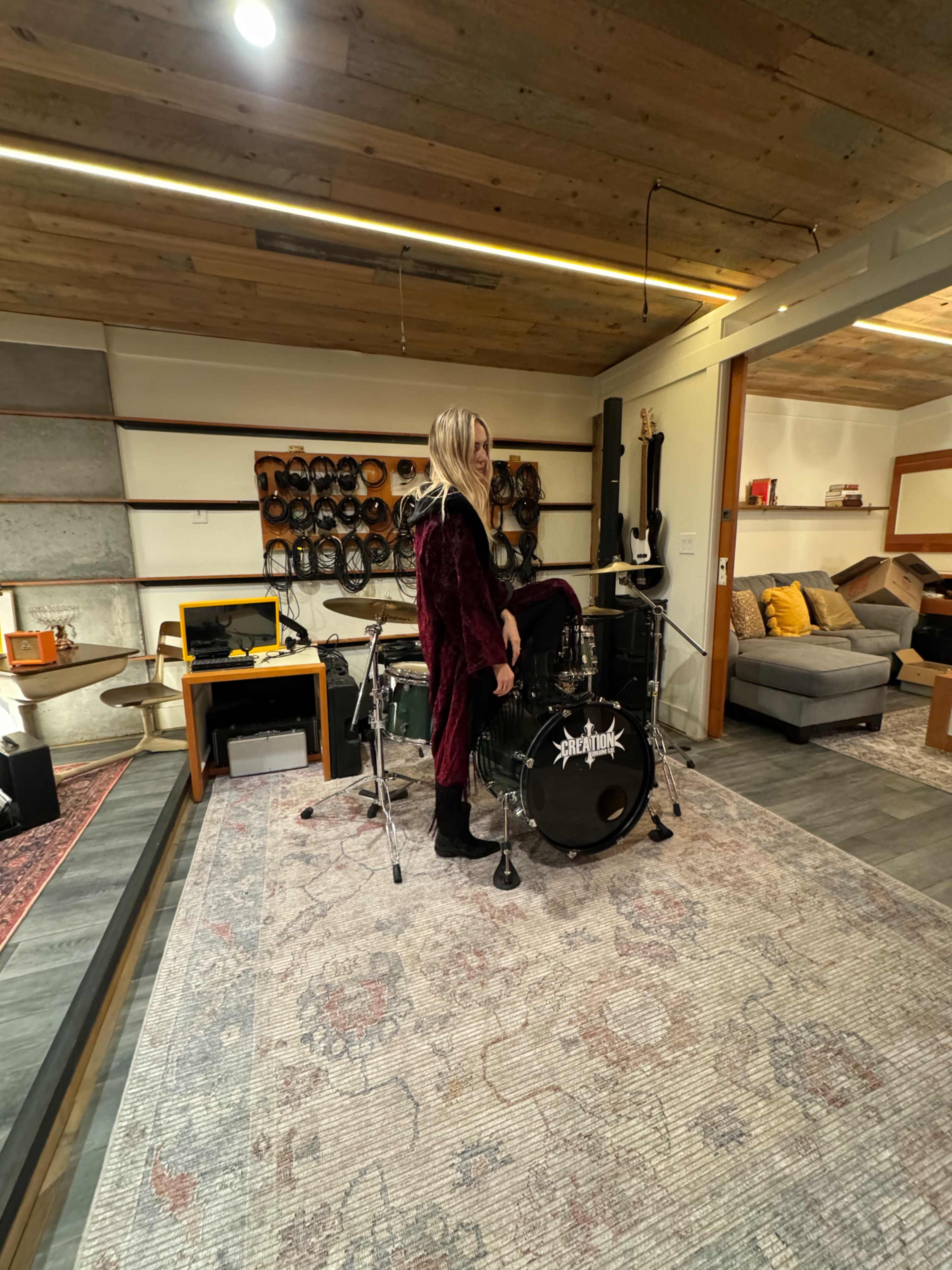 A person in a long, dark robe is standing on a drum set in a spacious room with wooden ceilings and various musical instruments displayed on the walls.