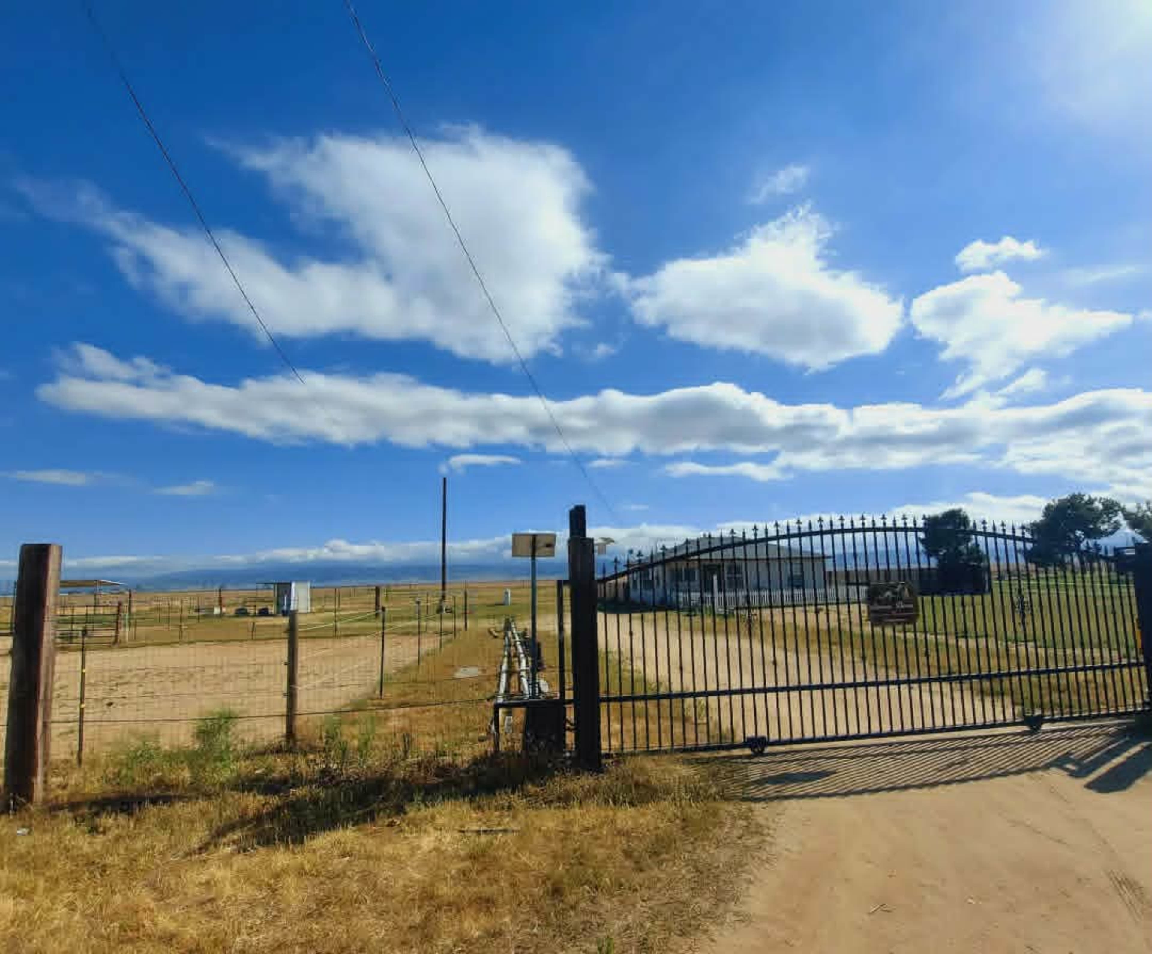 A metal gate leads into a wide open field under a blue sky with scattered clouds.
