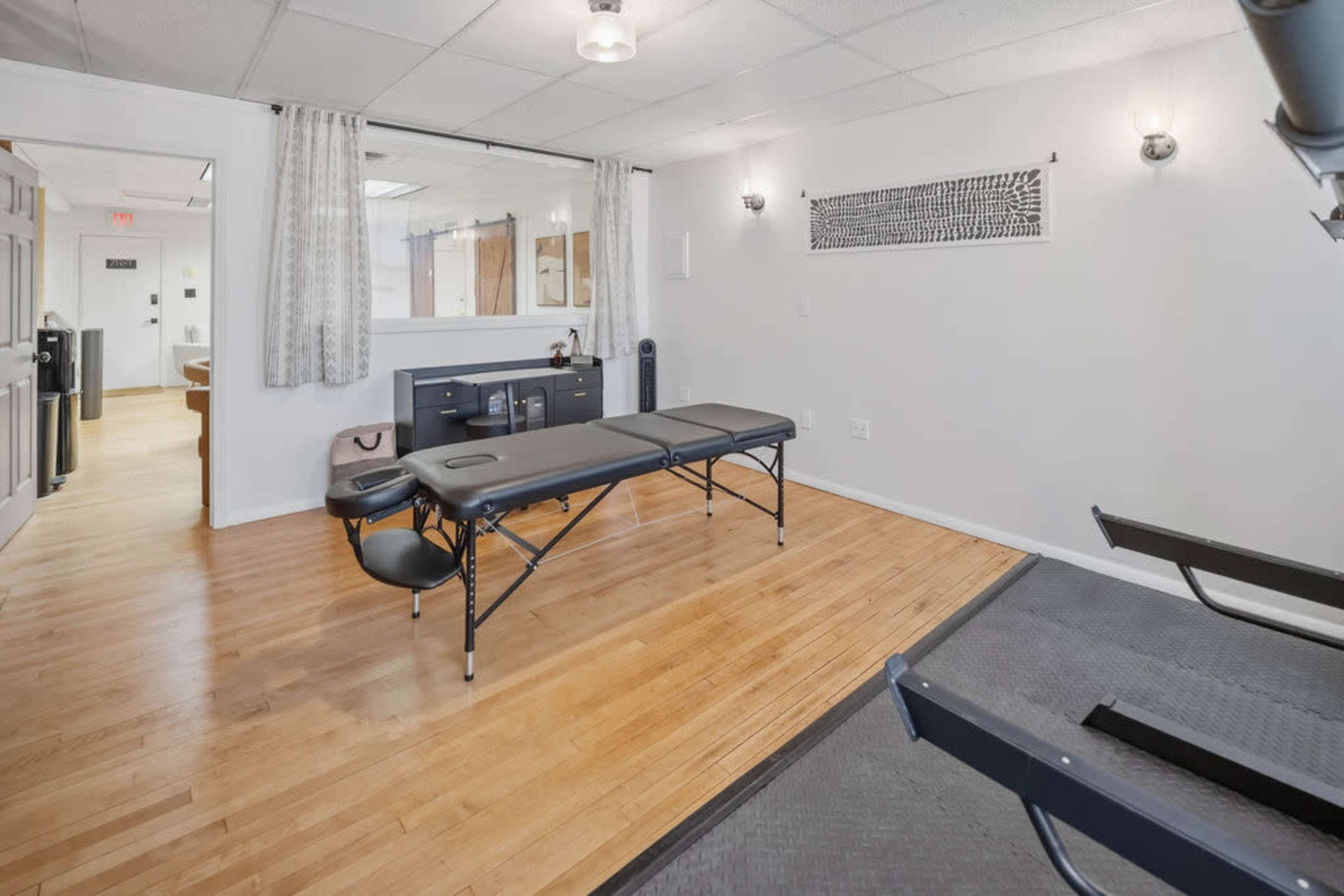 The image shows a clean, well-lit therapy room featuring a massage table, cabinetry, and neutral-colored walls with wooden flooring.