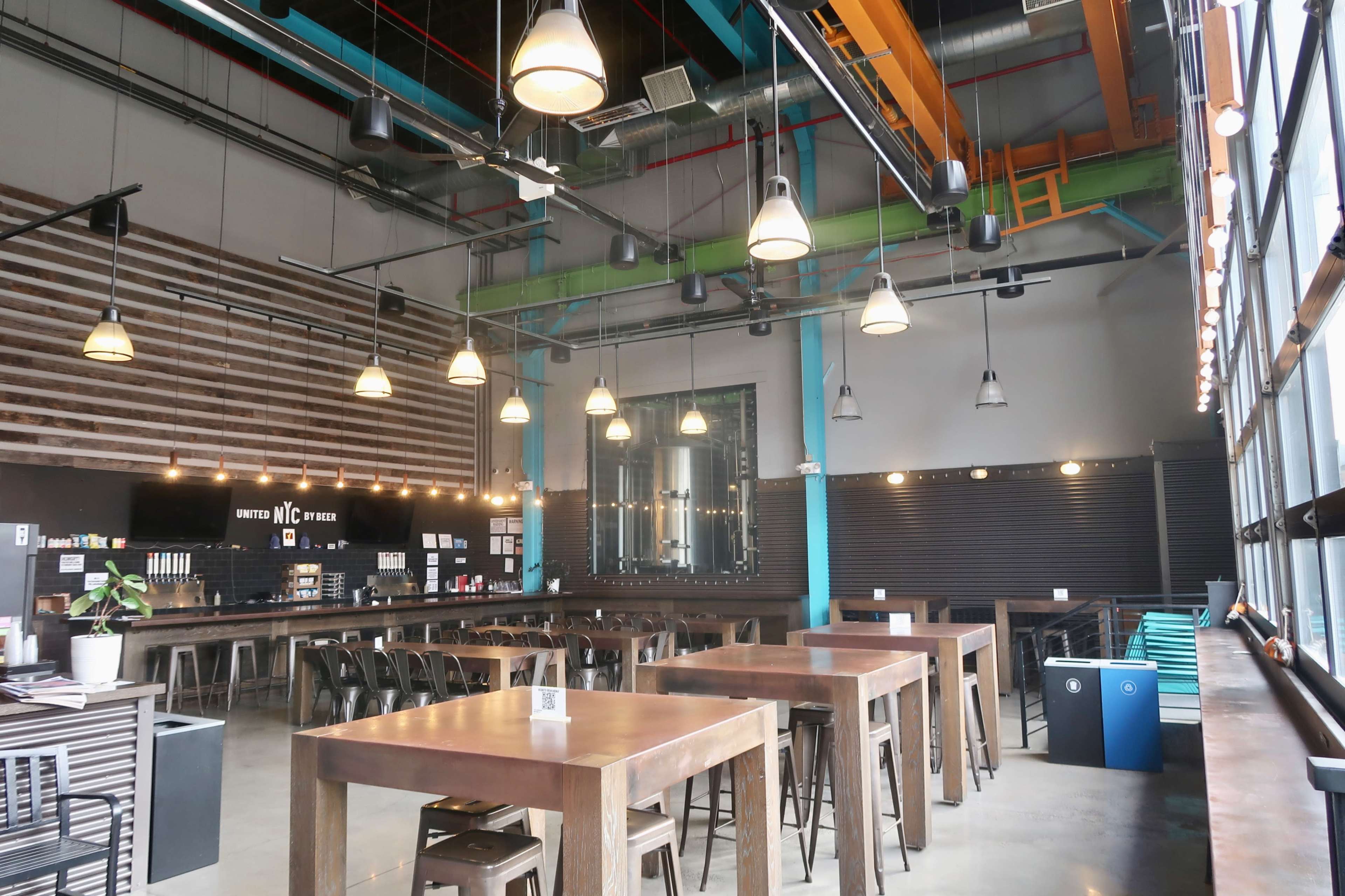 The image shows an industrial-style cafe interior featuring long wooden tables, pendant lighting, and a large window with seating arrangements against the walls.