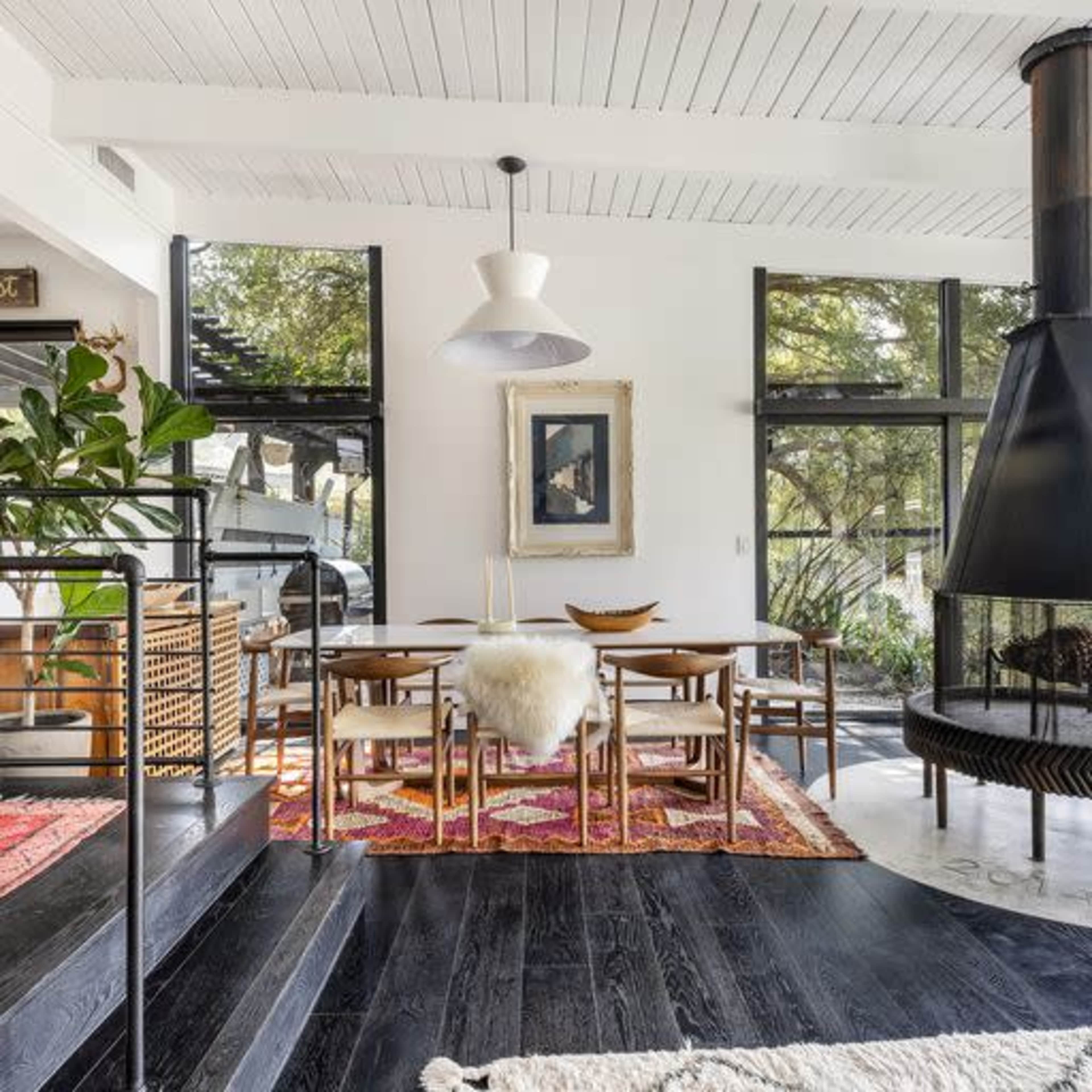 A modern interior with a dining area featuring a round table and wooden chairs, surrounded by large windows and a fireplace.