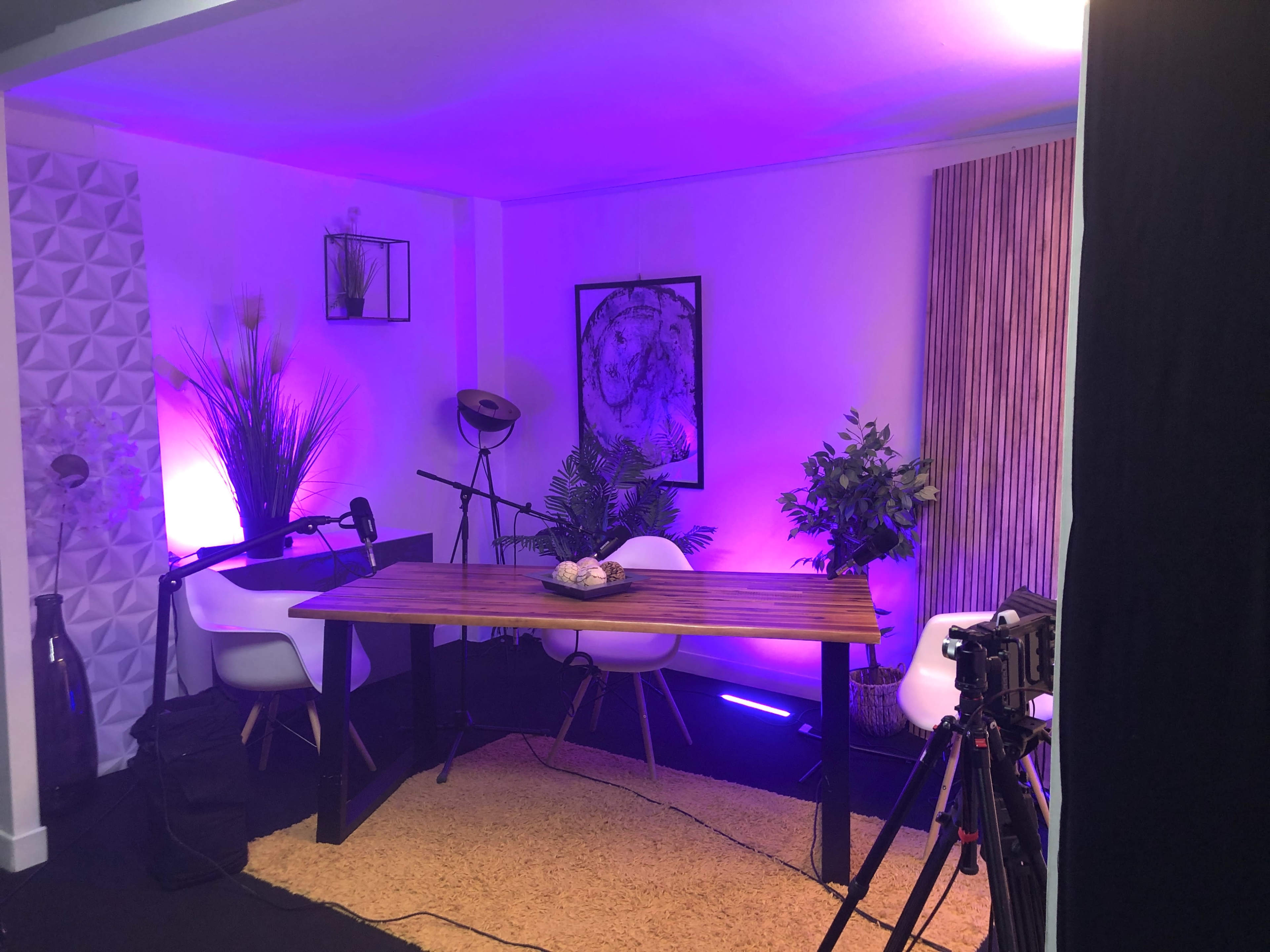 The image shows a modern studio setup with a wooden table, white chairs, and various decorative plants, illuminated by purple and white lighting.