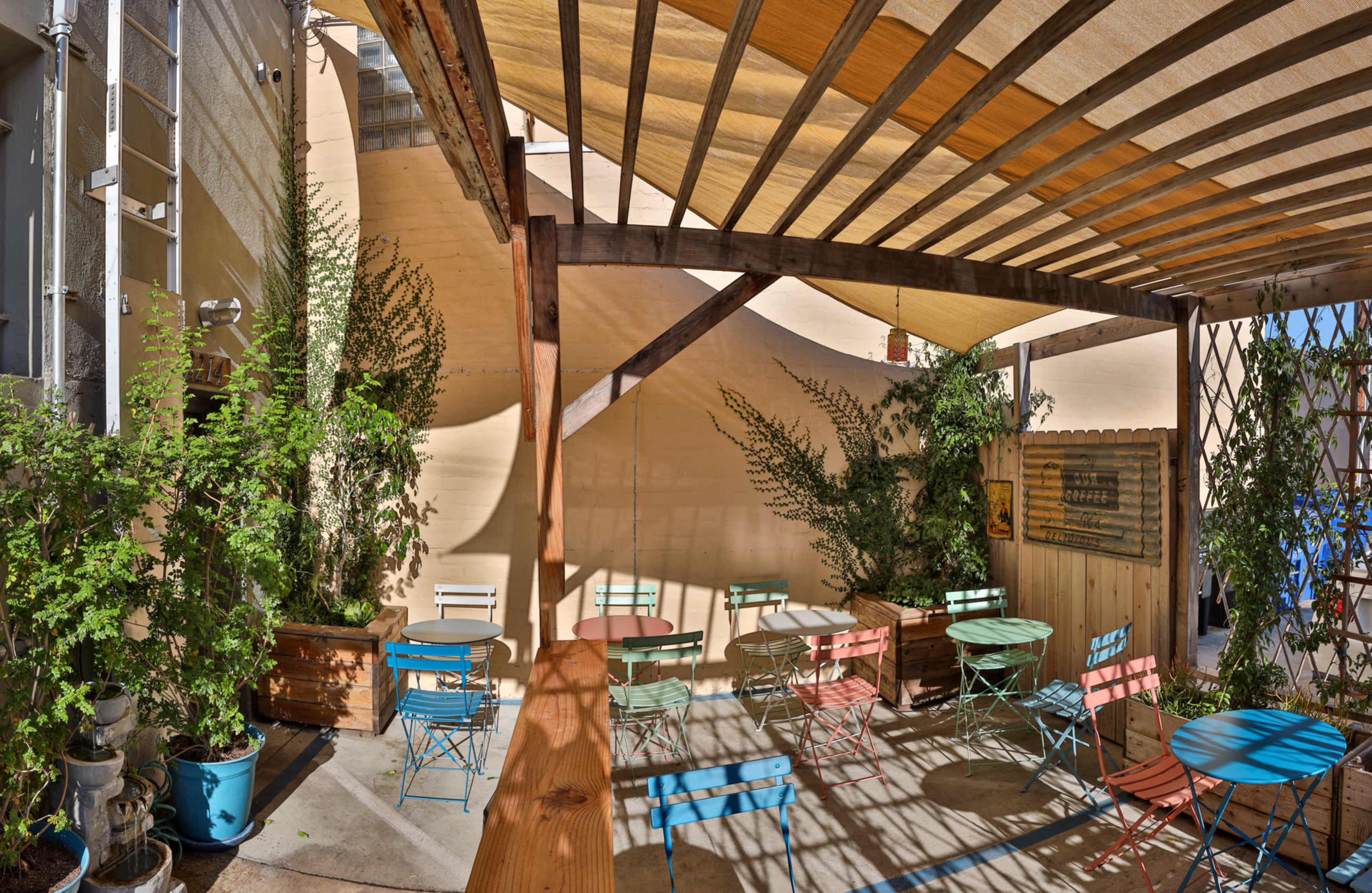 A patio area with colorful metal tables and chairs surrounded by greenery and shaded by a striped canopy.
