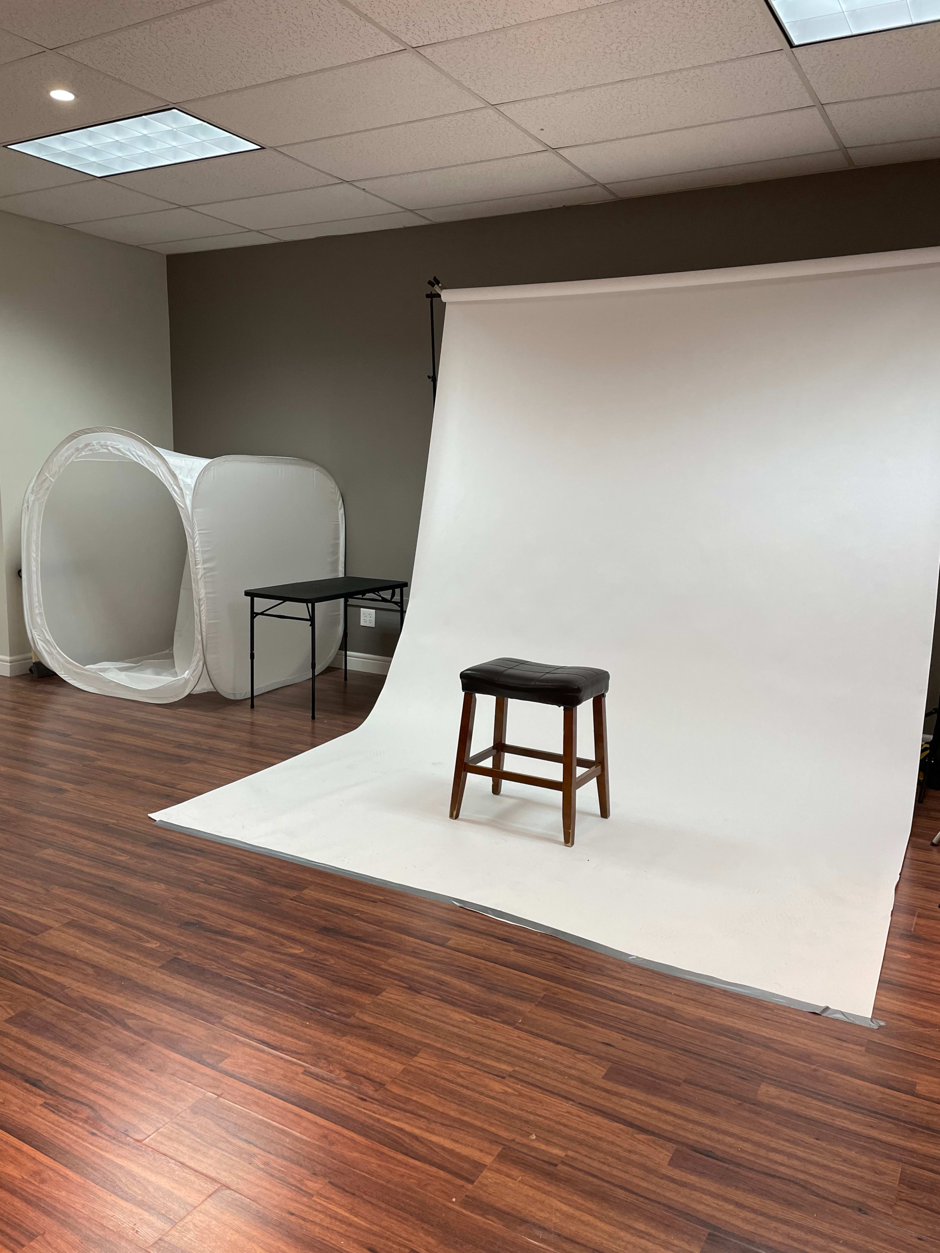 A photo studio setup with a white backdrop, a wooden stool, a folding table, and a light tent in the corner.