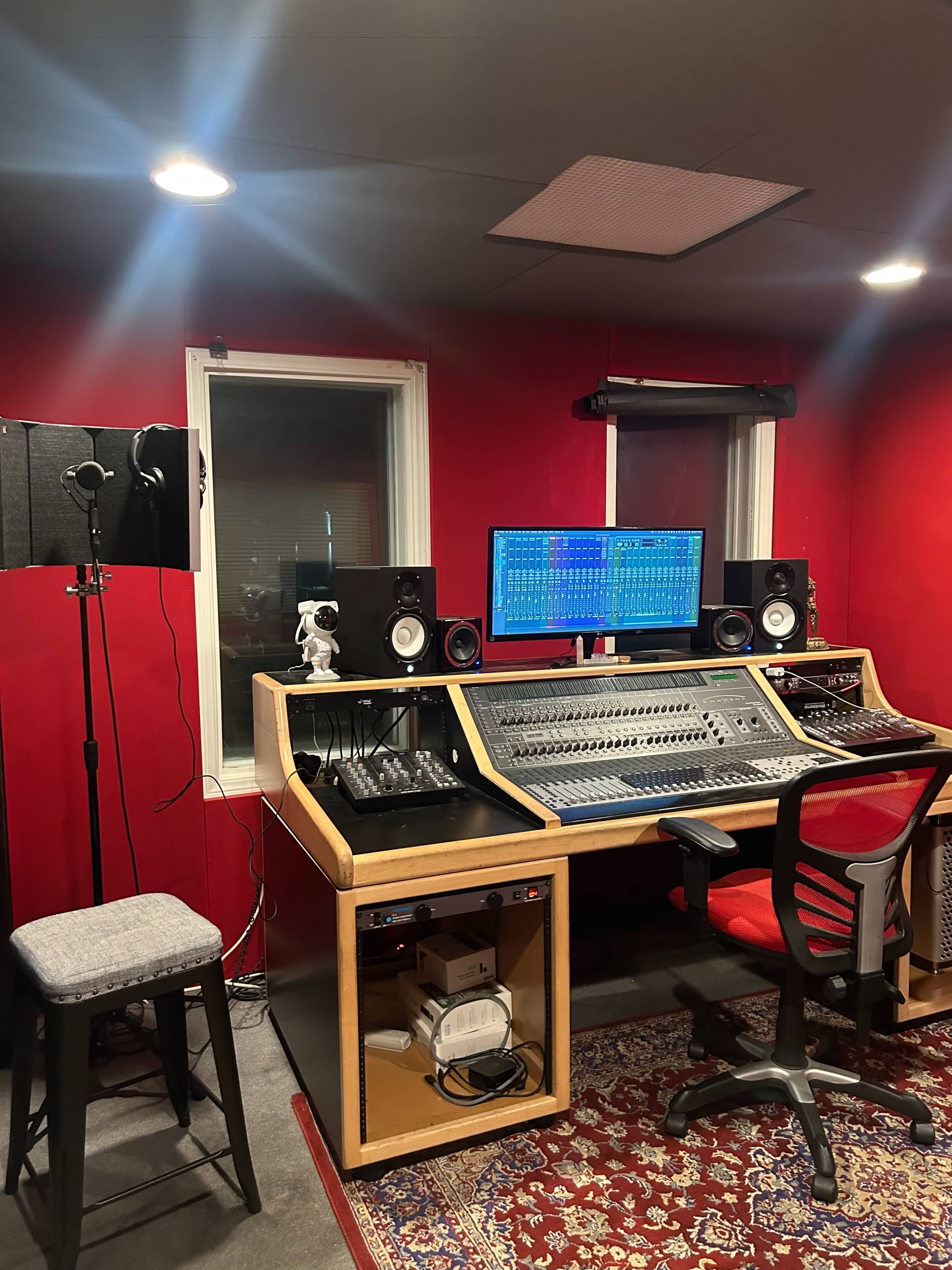 The image shows a music recording studio with a mixing console, computer monitors, speakers, and a microphone setup, all situated against red walls.