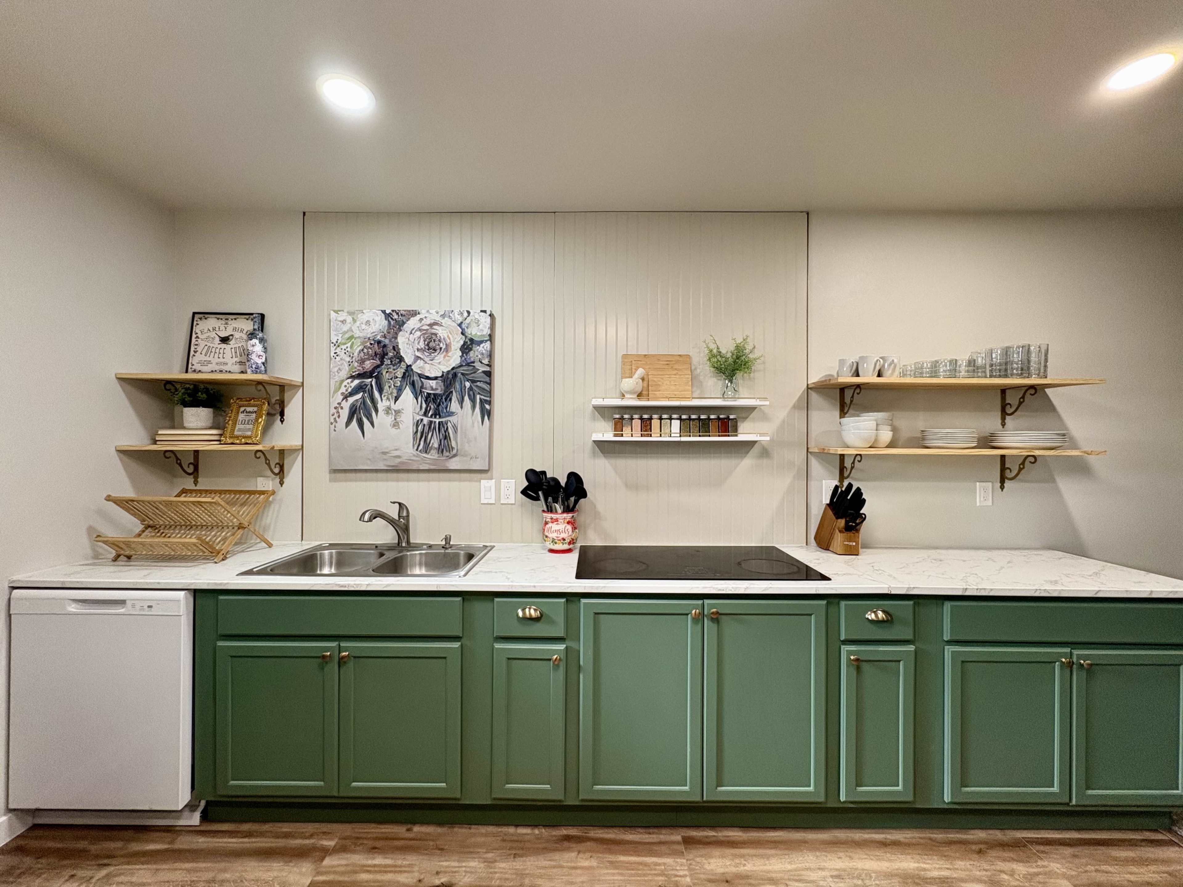A modern kitchen features green cabinetry, a double sink, a countertop with a stovetop, and floating shelves displaying decorative items and kitchenware.