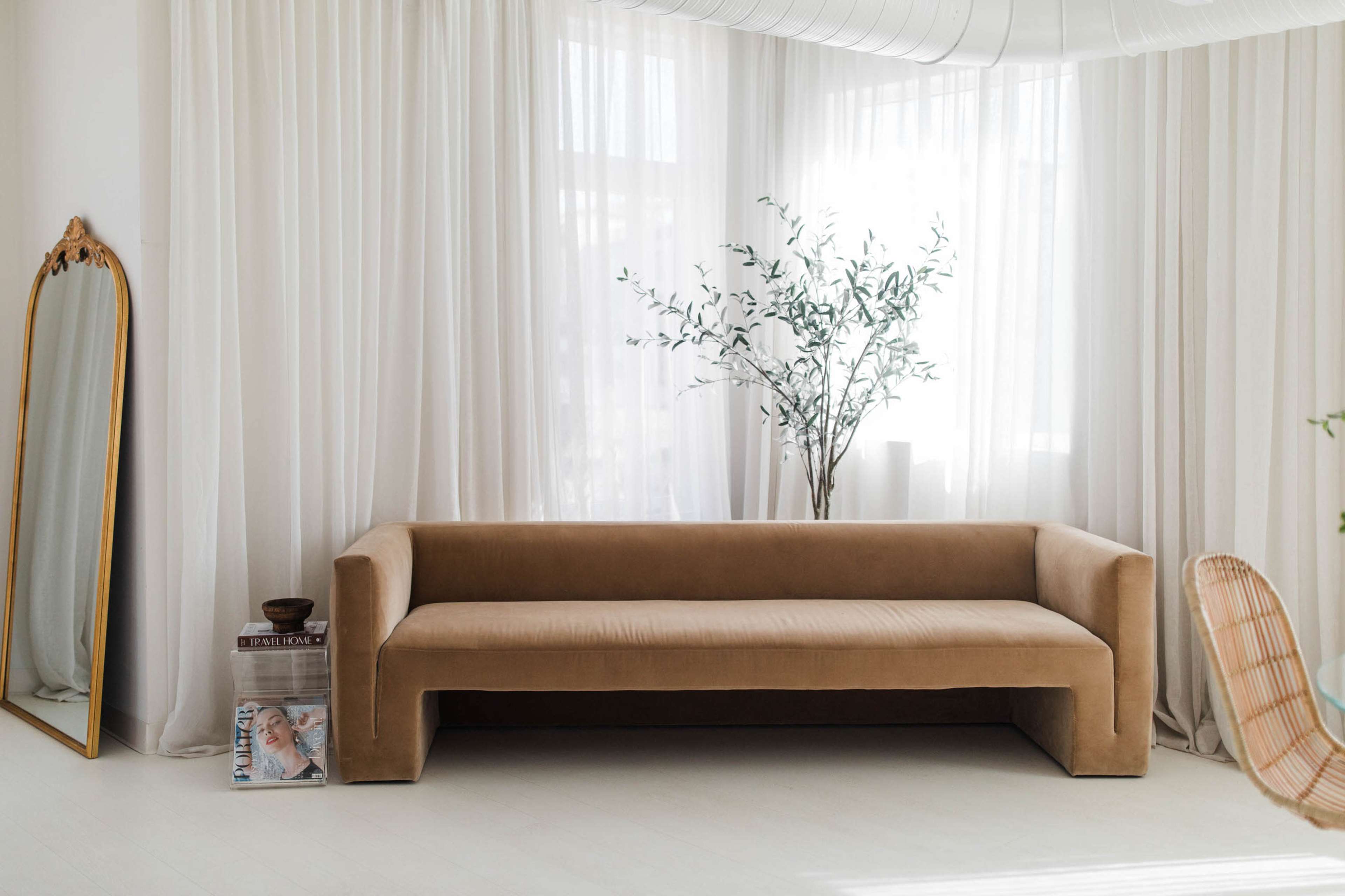A minimalist living room features a beige velvet sofa, a large mirror, a decorative plant, and white sheer curtains.