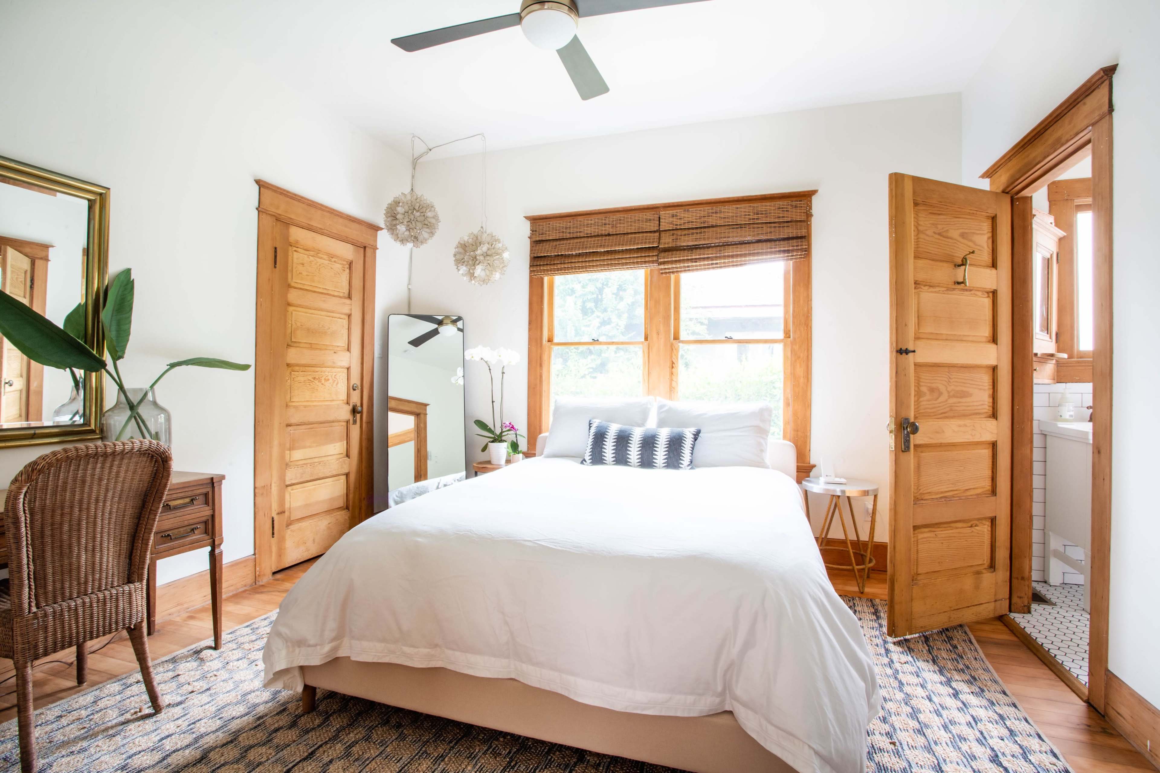 A well-lit bedroom featuring a double bed, wooden furniture, a plant, and large windows with bamboo shades.