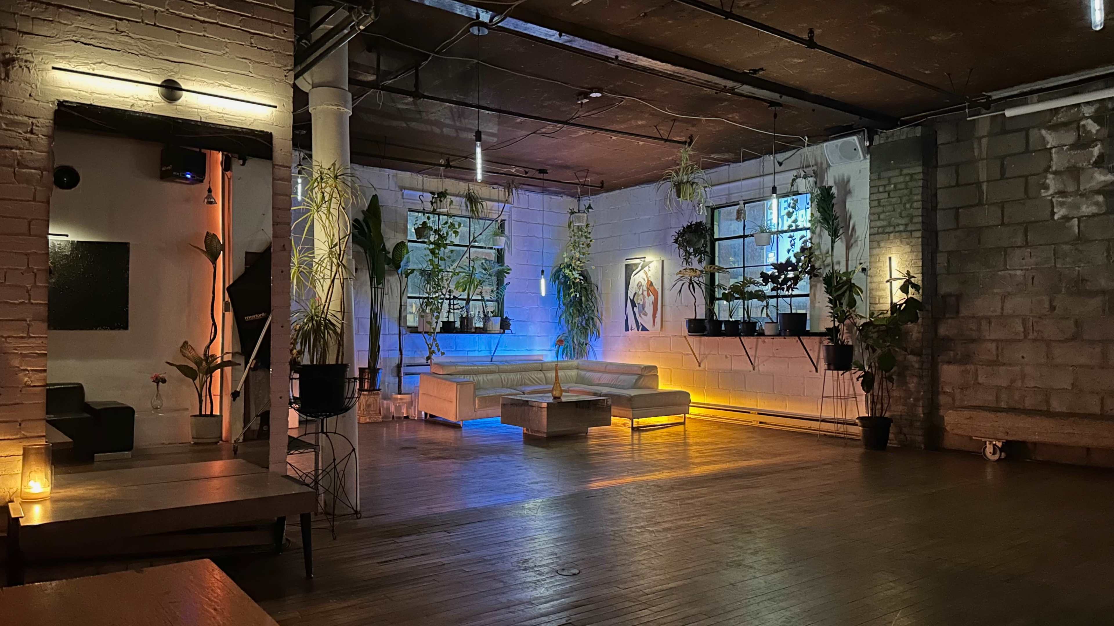 The image shows a spacious indoor lounge area with plants, warm lighting, and a modern seating arrangement.