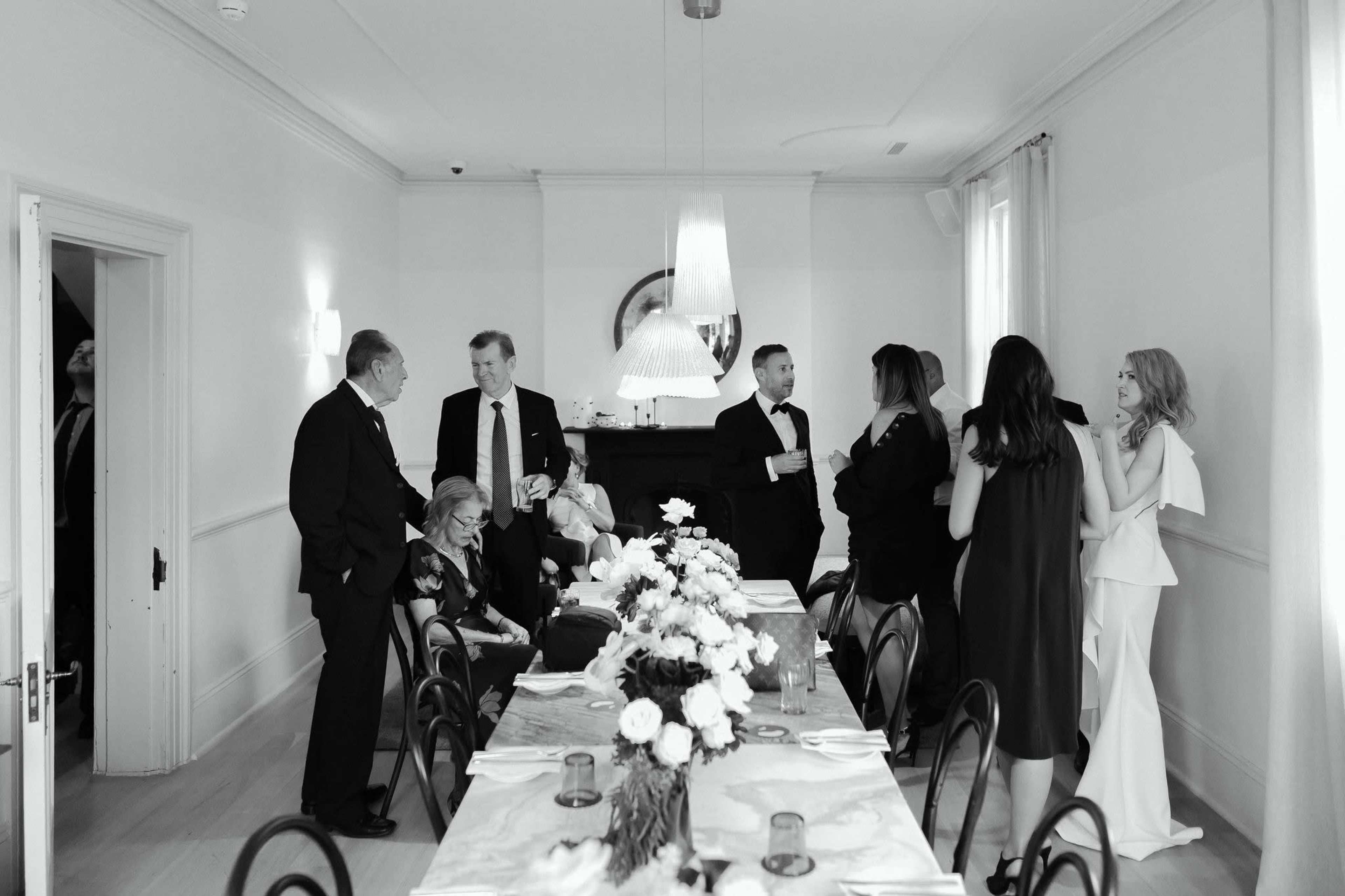 A group of elegantly dressed individuals socializes around a dining table adorned with flowers in a well-lit room.