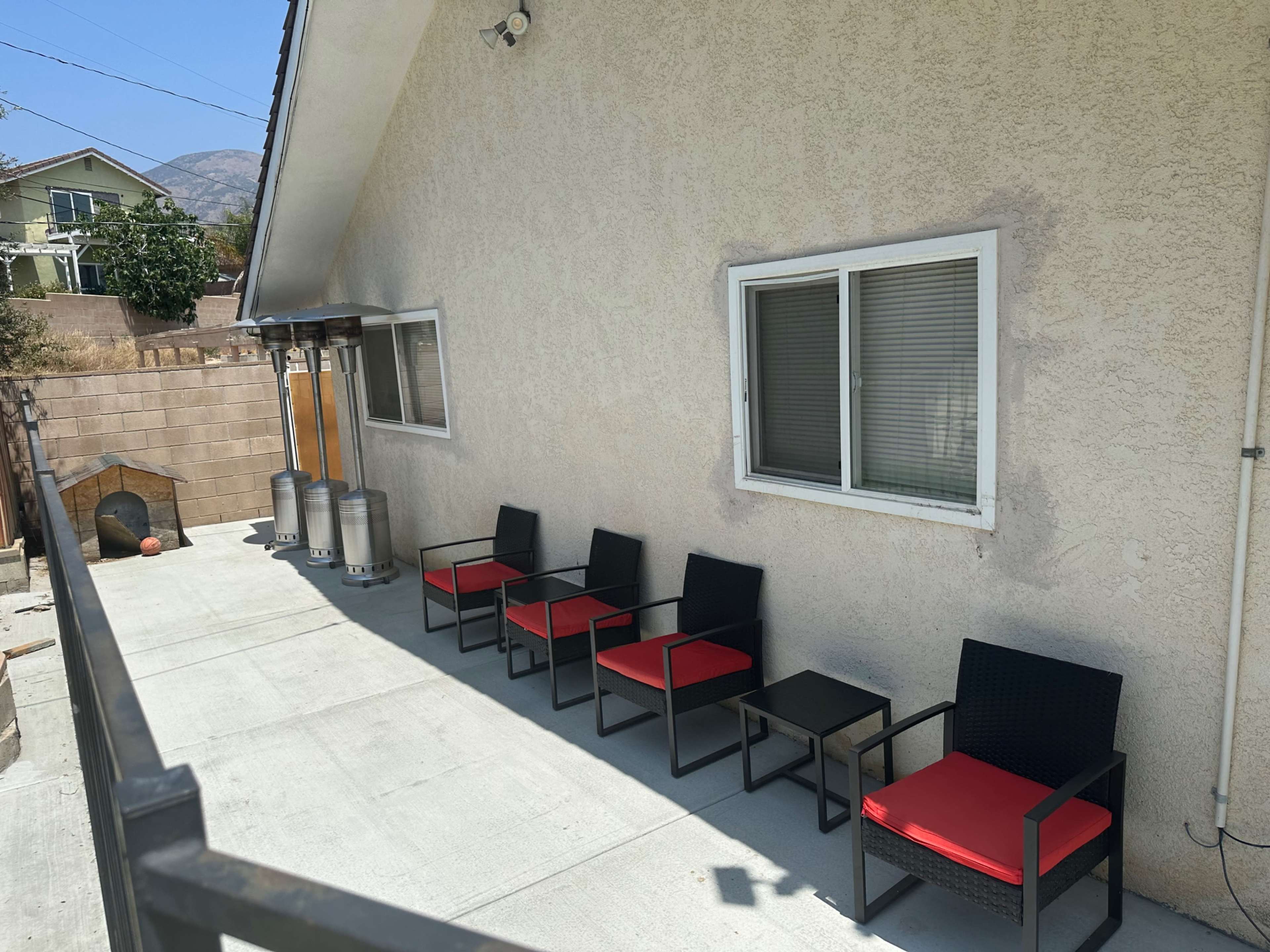 A backyard patio with five black chairs featuring red cushions, a small table, and two patio heaters against a tan house wall.