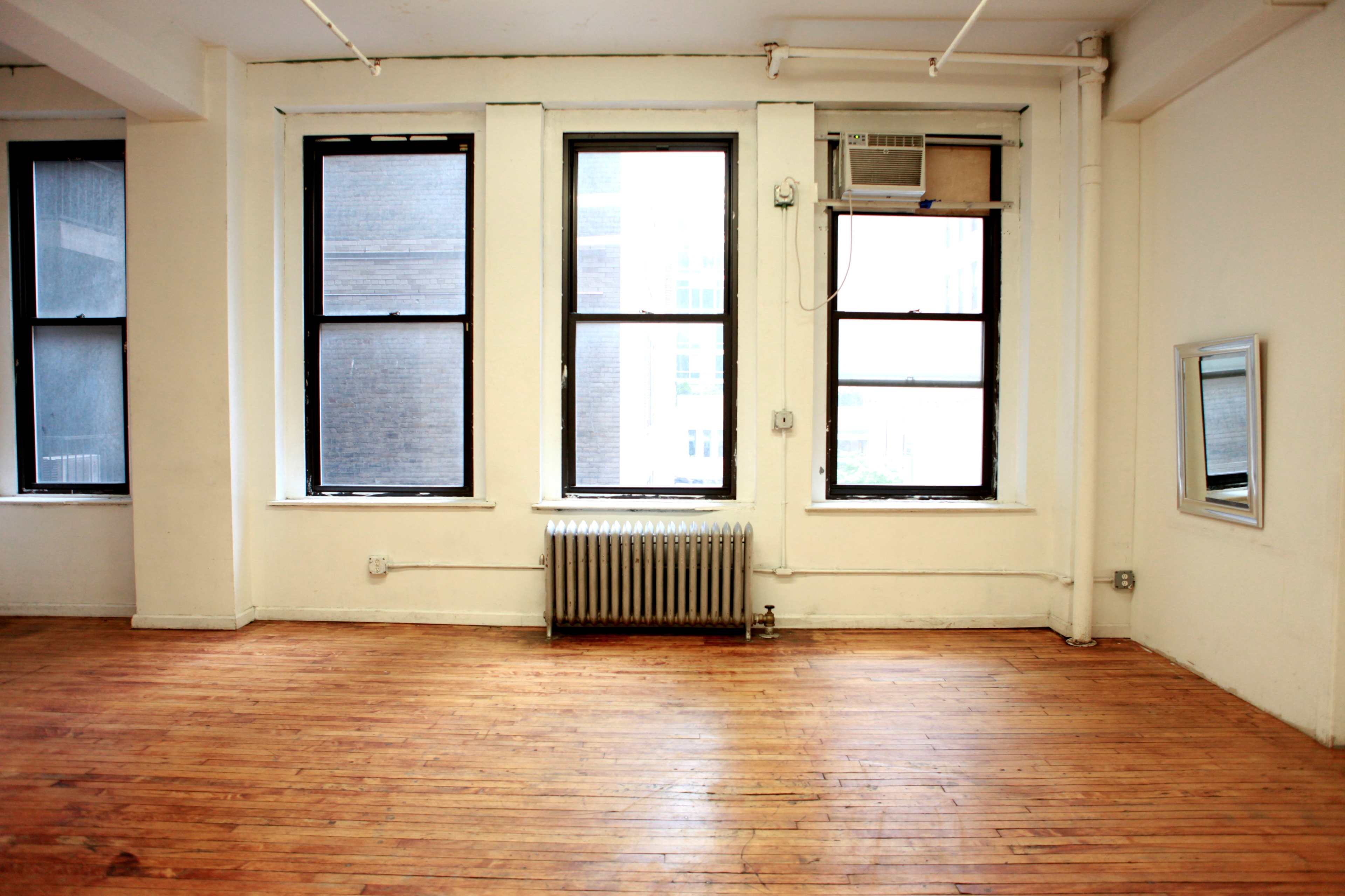 A vacant room with hardwood floors, large windows, an air conditioning unit, and a wall mirror.