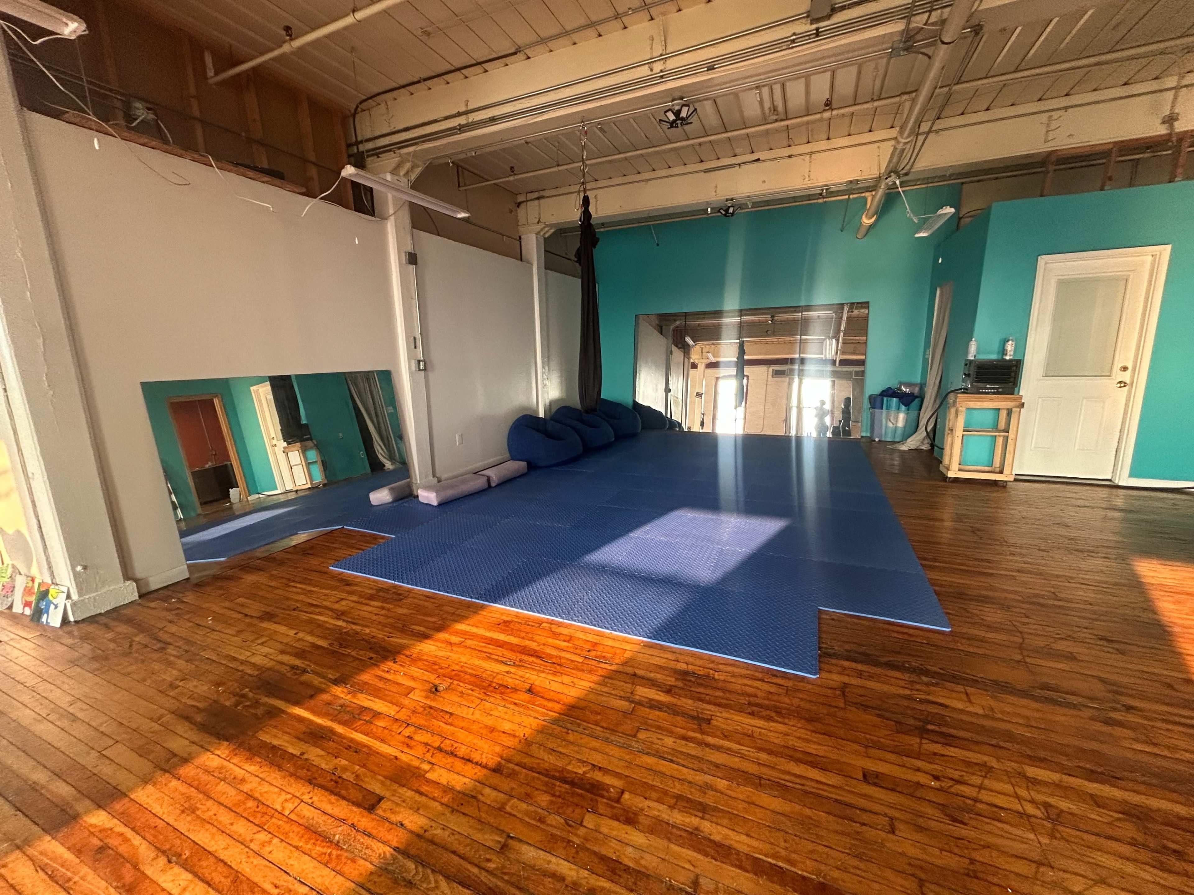 The image shows a spacious room with wooden floors, blue mats laid out on a portion of the floor, and mirrors on one wall.