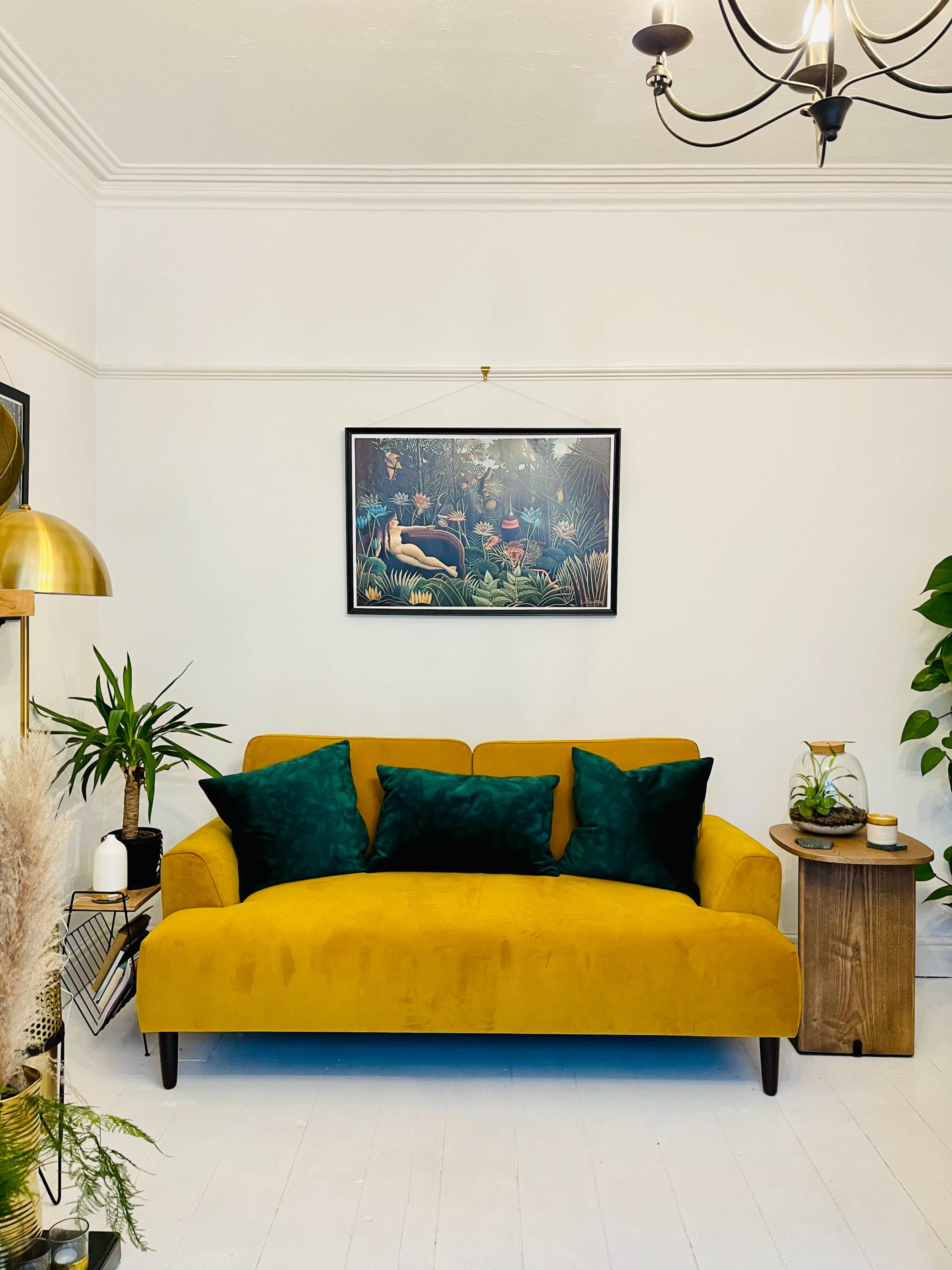 A mustard yellow couch with green cushions sits against a white wall, adorned with a framed artwork above it, complemented by decorative plants and a side table.