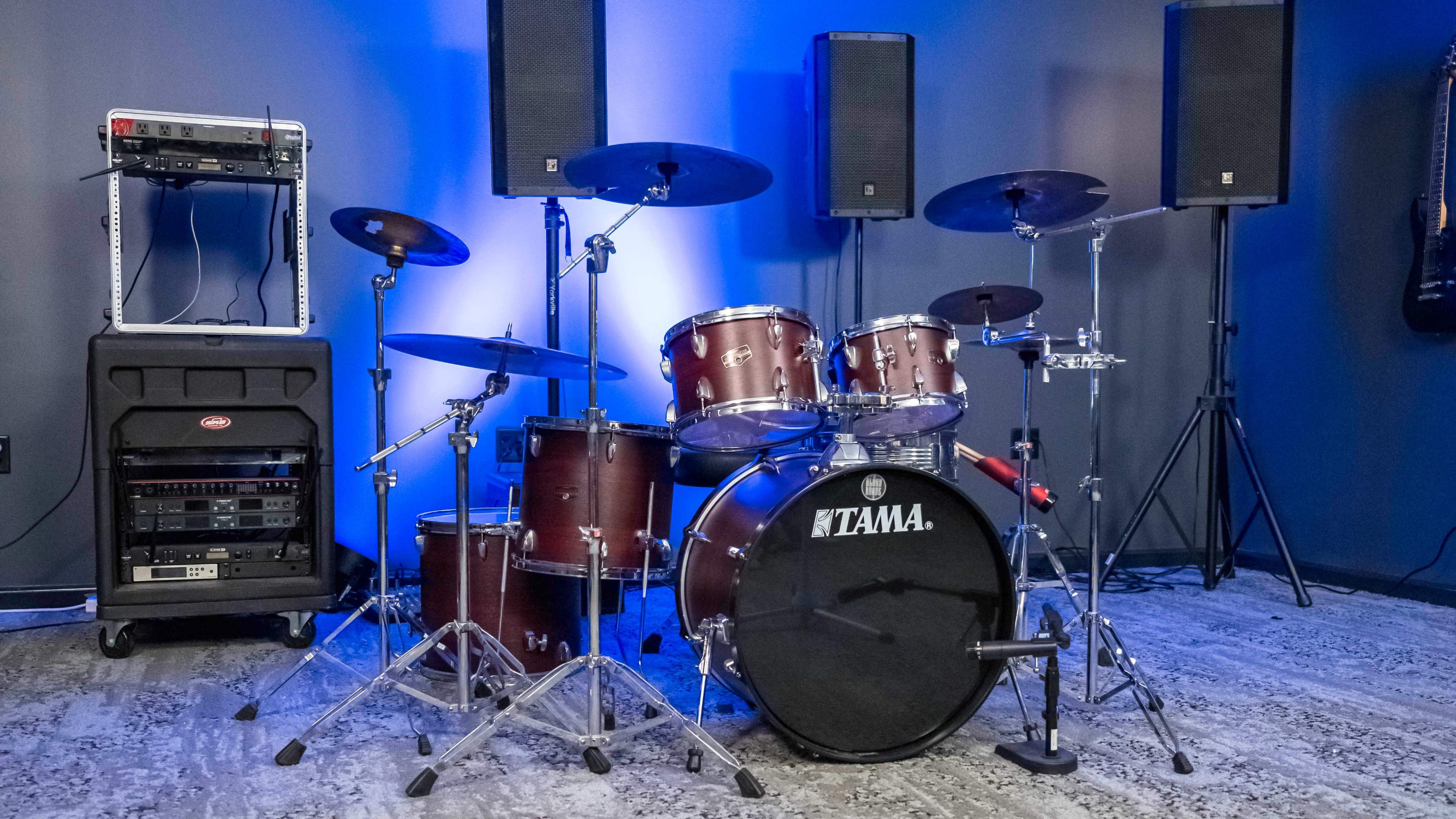 A drum set is positioned in a room with blue lighting, flanked by large speakers and audio equipment.
