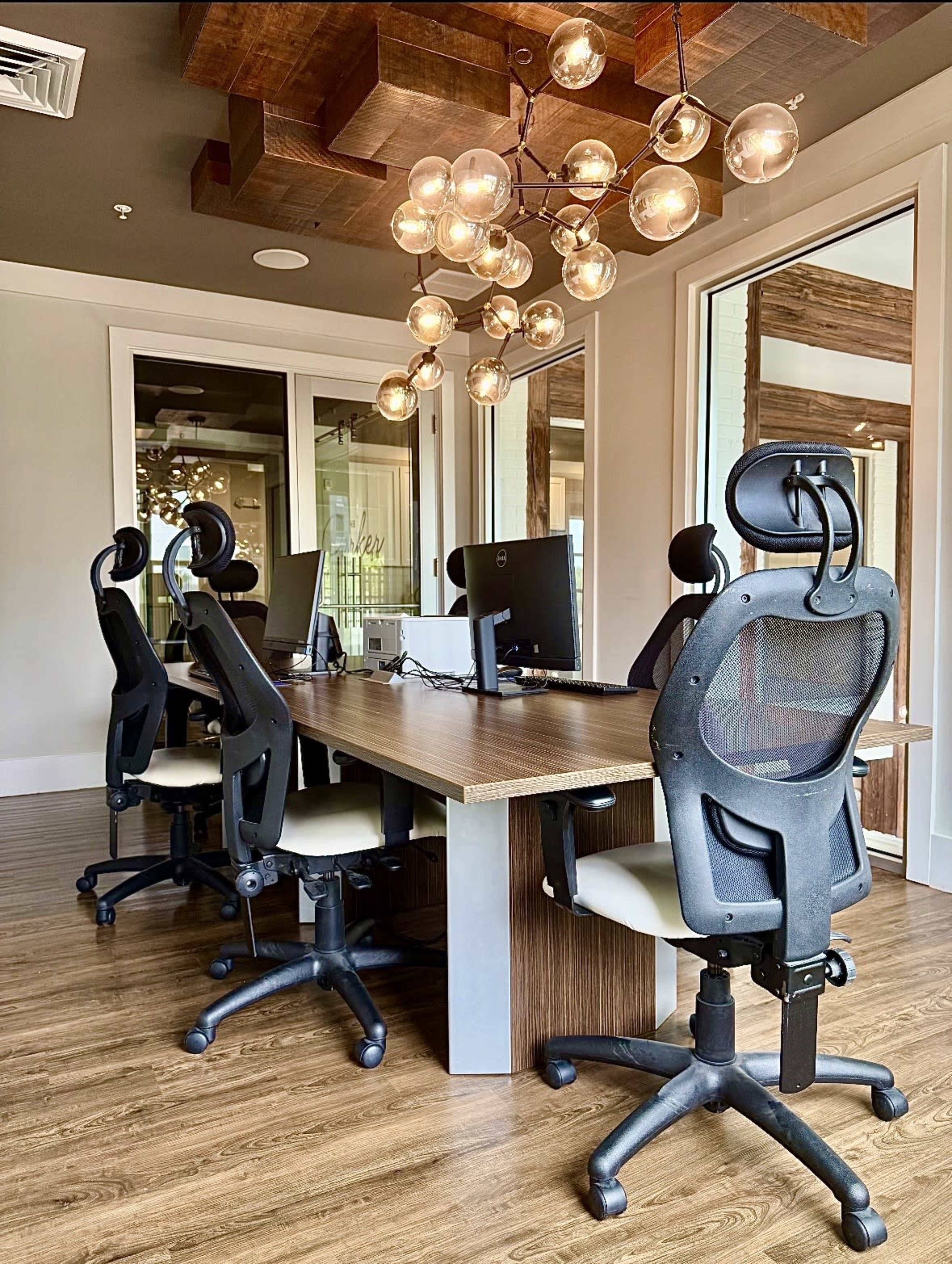 The image shows a modern office space featuring a large wooden table surrounded by four black office chairs, with a decorative light fixture hanging above.