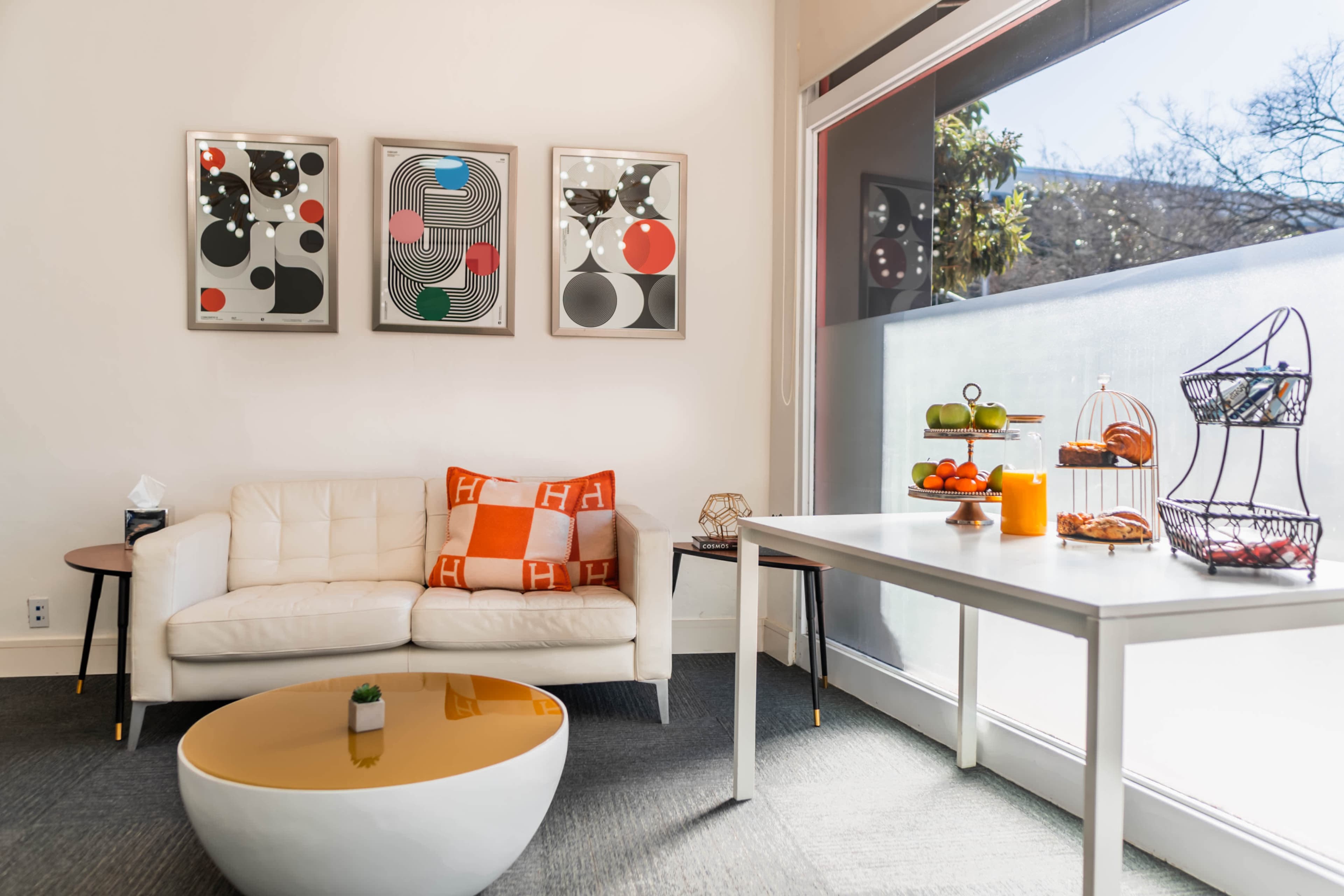 A modern living room features a white couch, a round coffee table, three framed abstract artworks, and a dining table with decorative fruits.