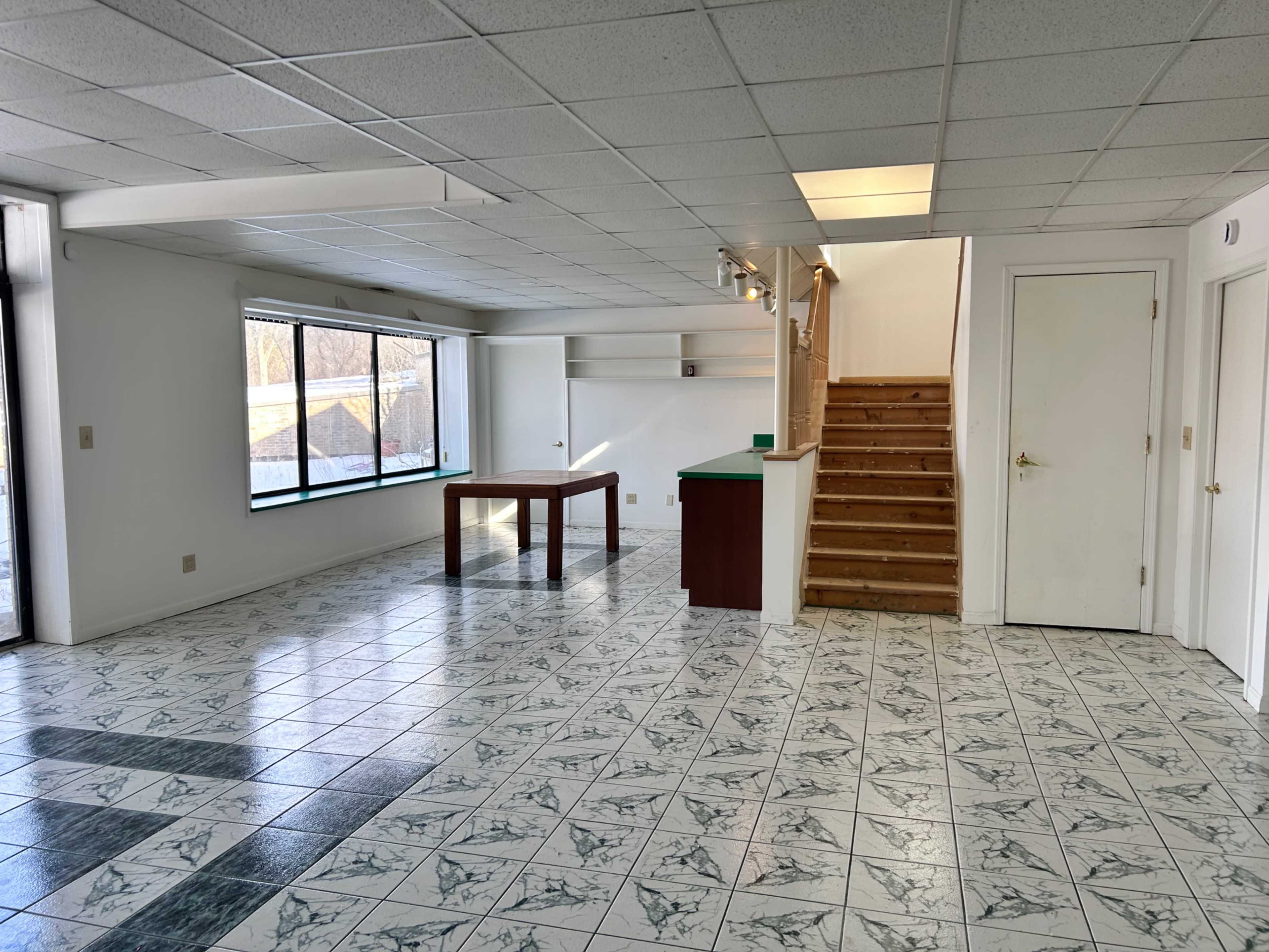 The image shows a spacious room with tiled flooring, large windows, a wooden table in the center, a small green counter, and a staircase leading to another level.