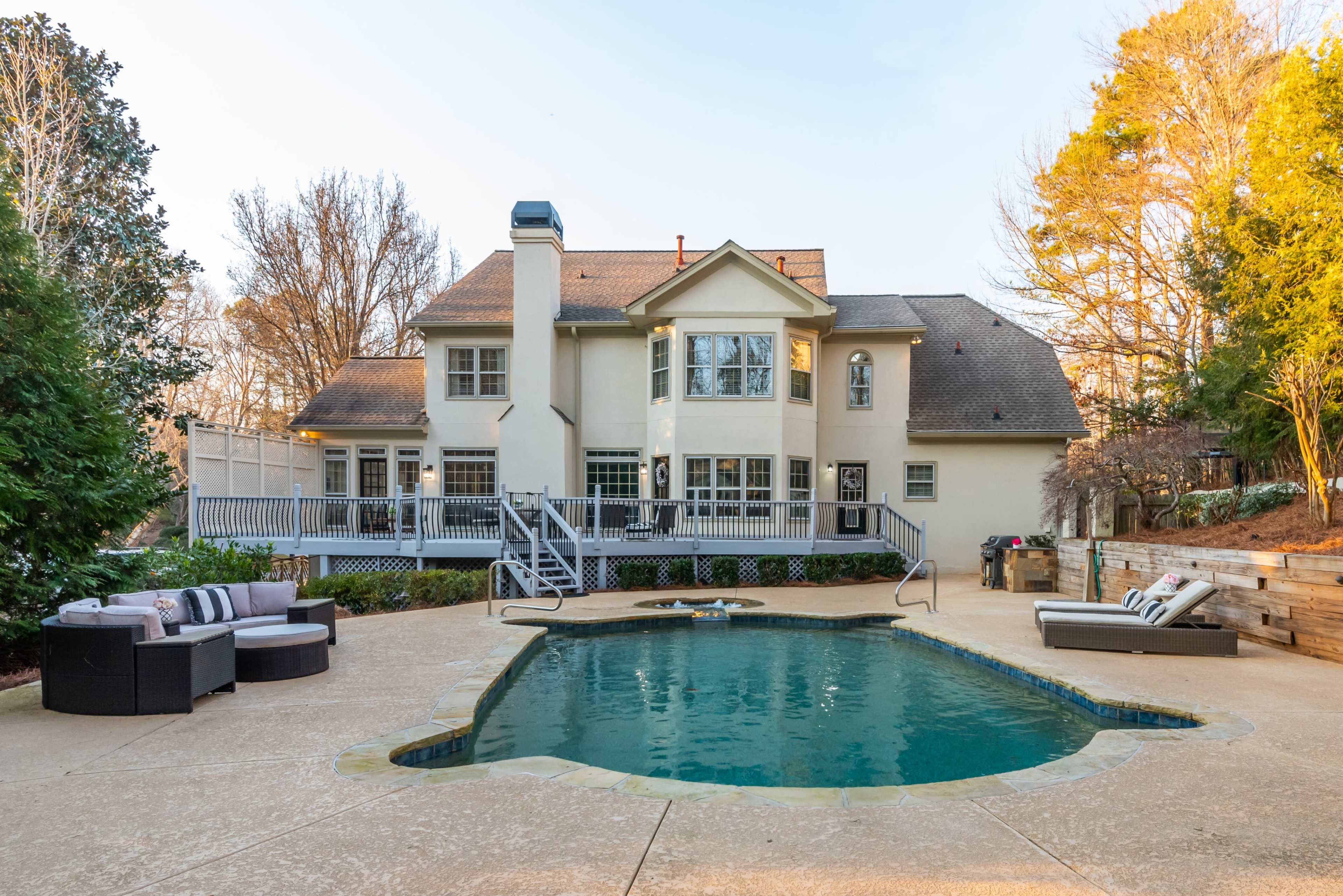 A large house with multiple windows sits behind a swimming pool and patio area, featuring outdoor seating and landscaped surroundings.