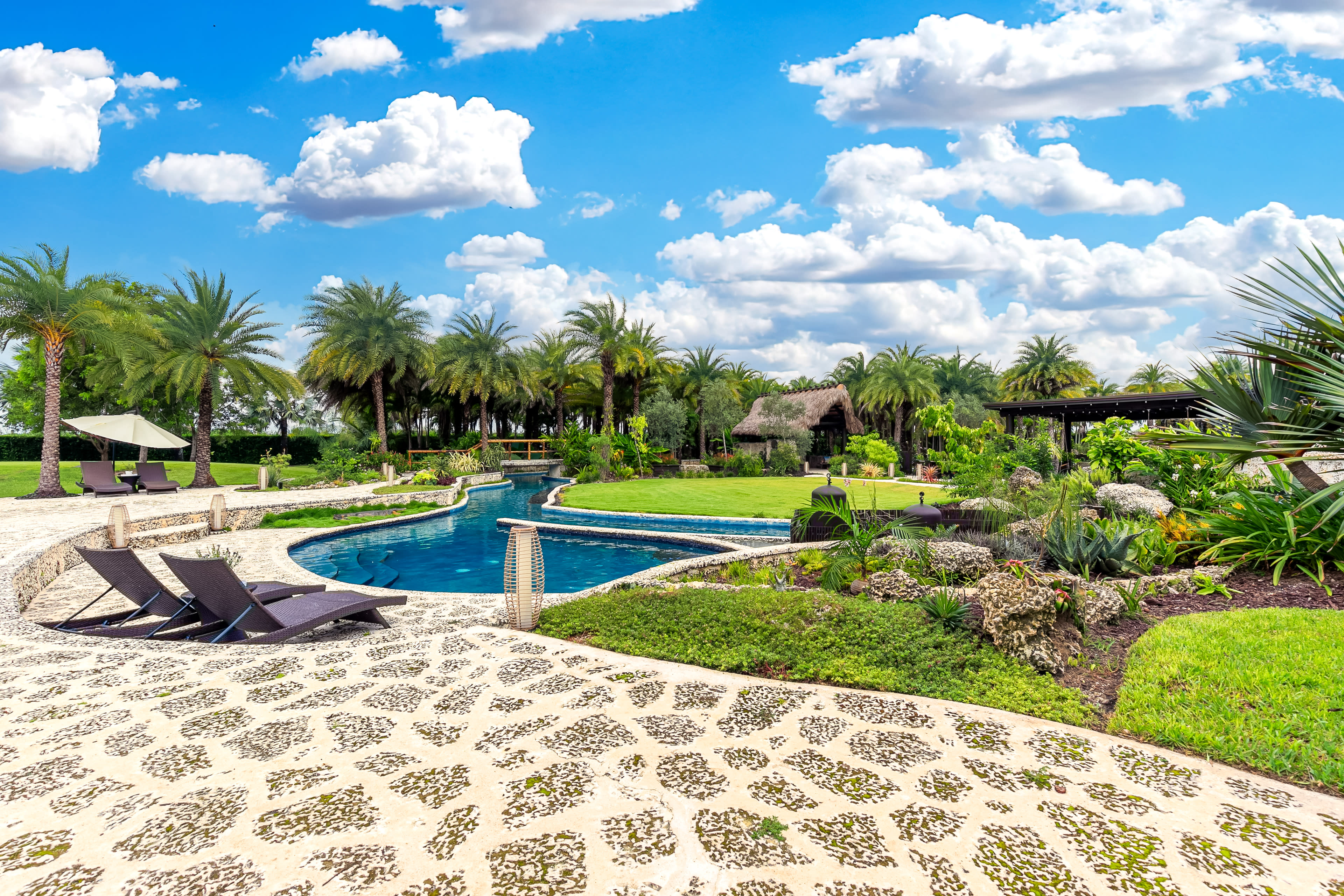 A curved swimming pool surrounded by palm trees and stone pathways is positioned within a landscaped yard.