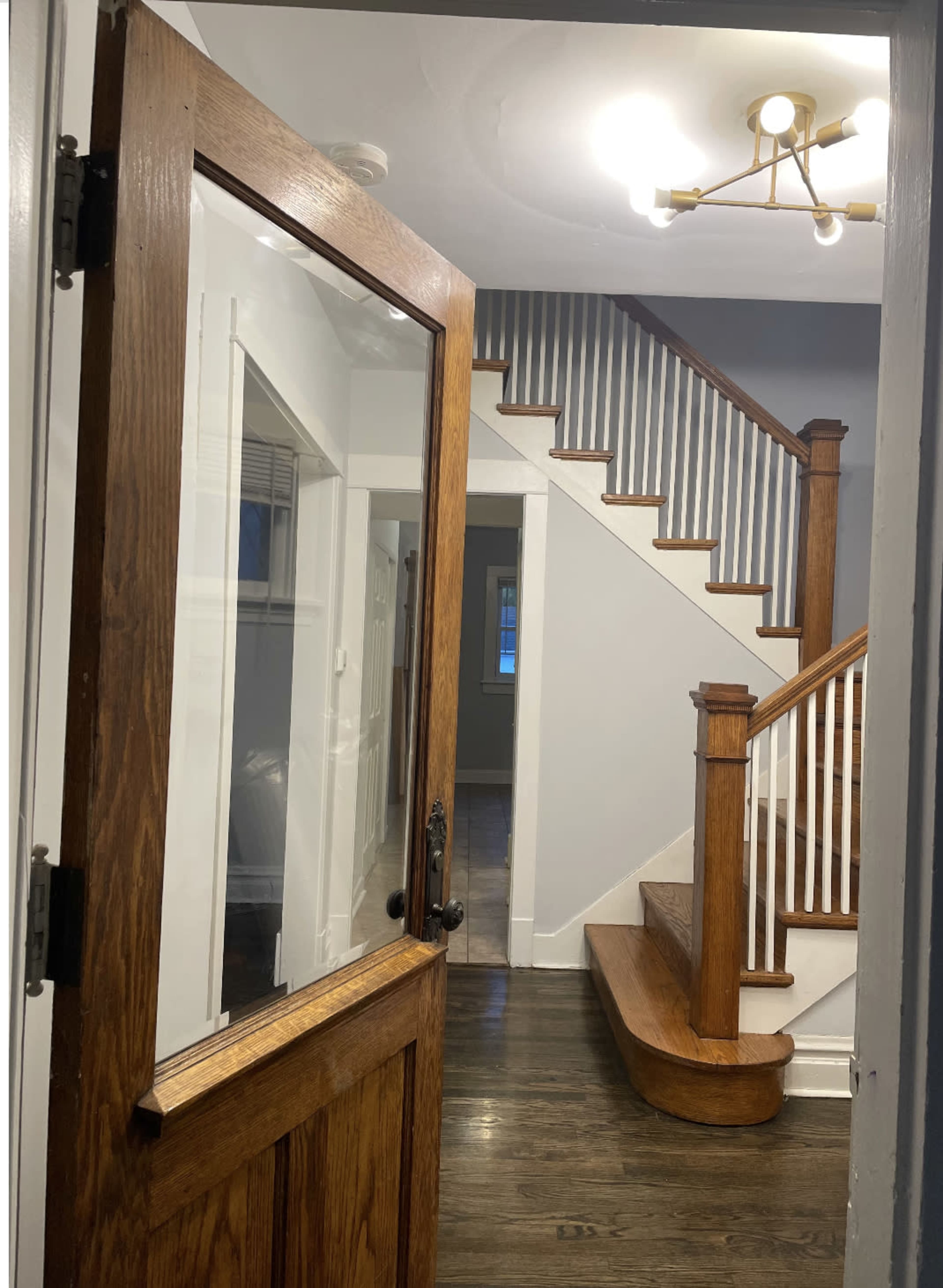A wooden door opens to reveal a staircase with wooden railings and a light fixture above, leading into a hallway.