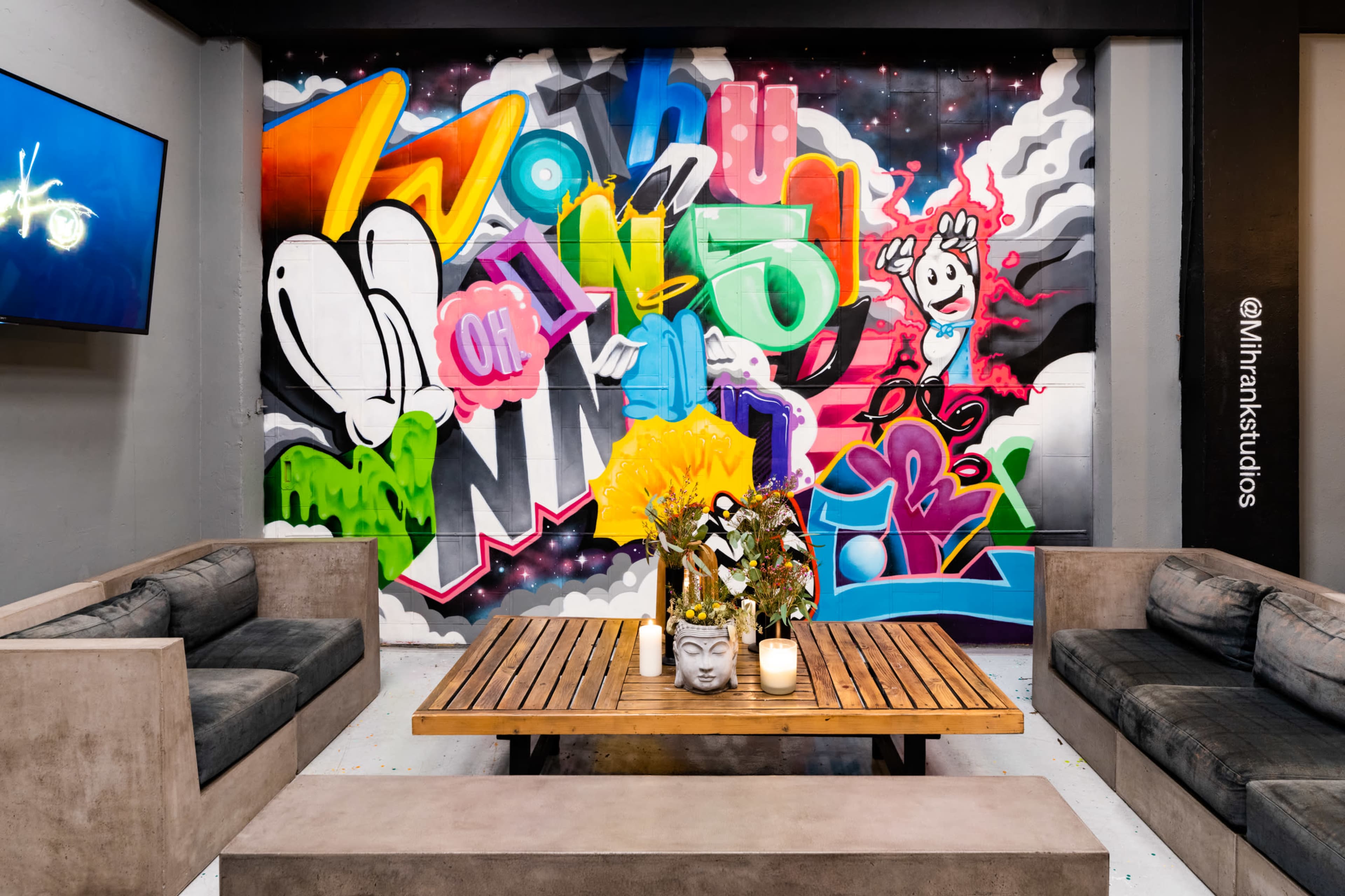 The image shows a stylish lounge area with a wooden coffee table at the center, surrounded by gray sofas, and a vibrant graffiti wall as the backdrop.