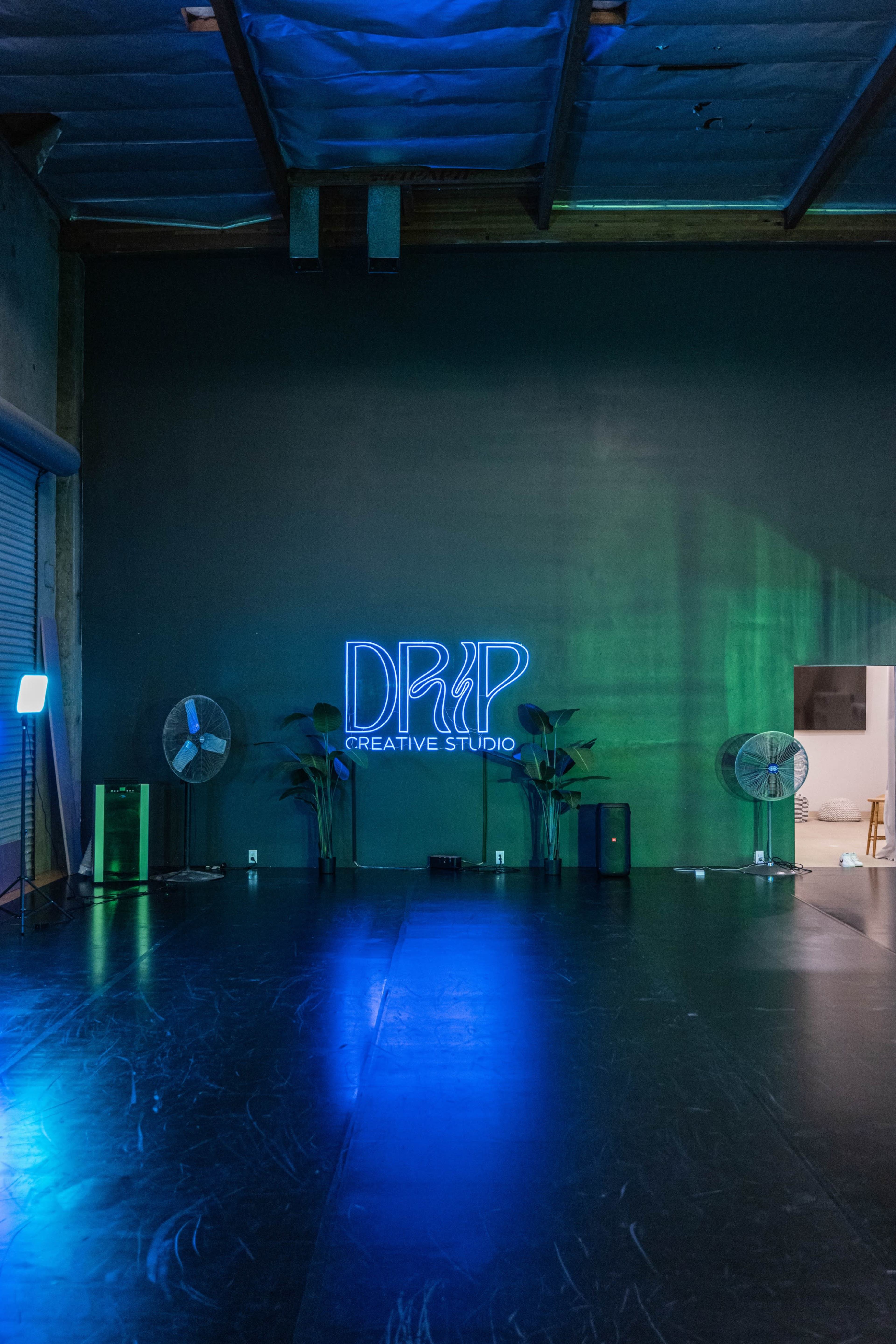 A spacious studio interior featuring a neon sign that reads "DRIP CREATIVE STUDIO" on a dark wall, accompanied by plants and standing fans.