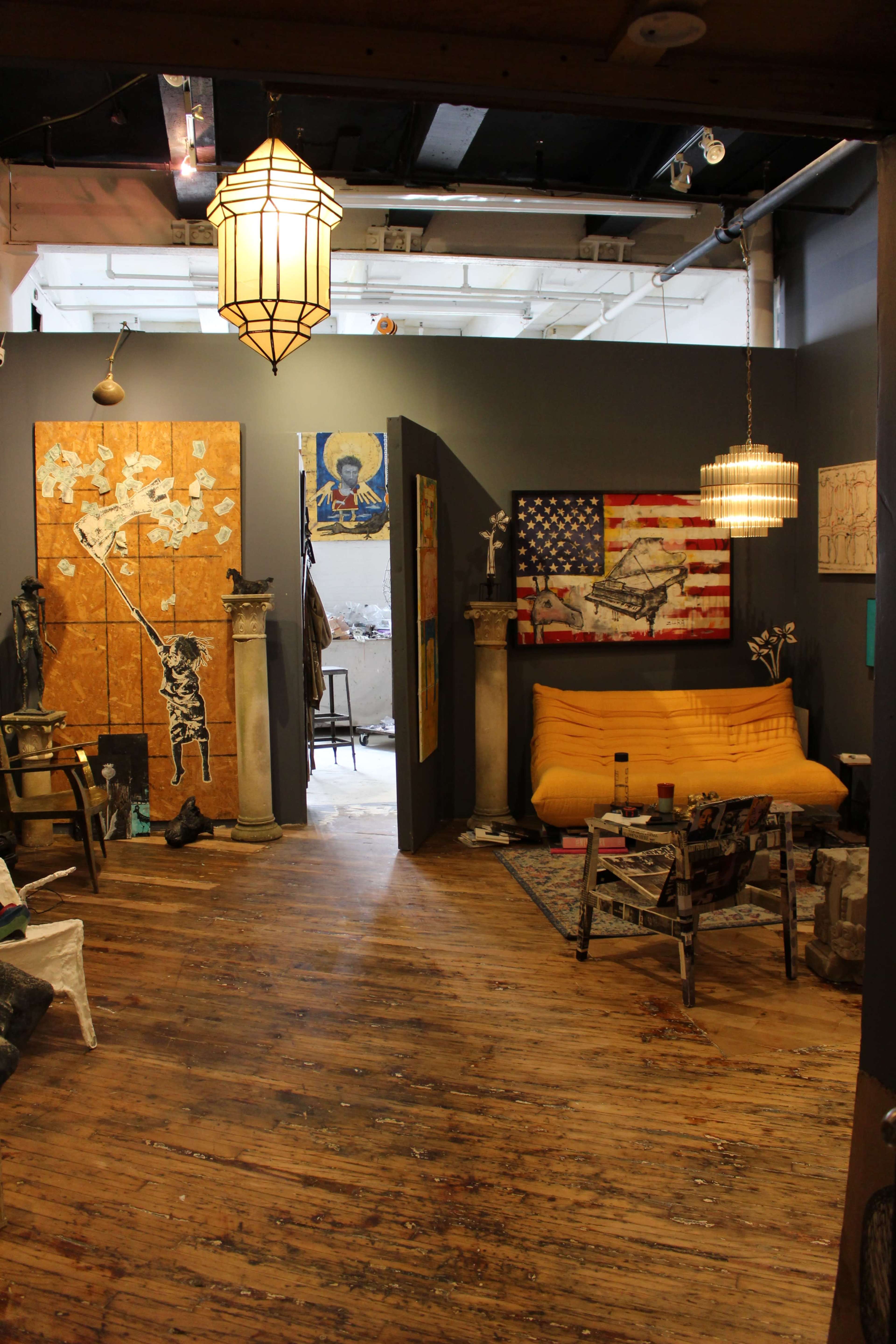 The image shows an artist's studio featuring wooden floors, an orange couch, various artworks on the walls, and a partially visible workspace in the background.