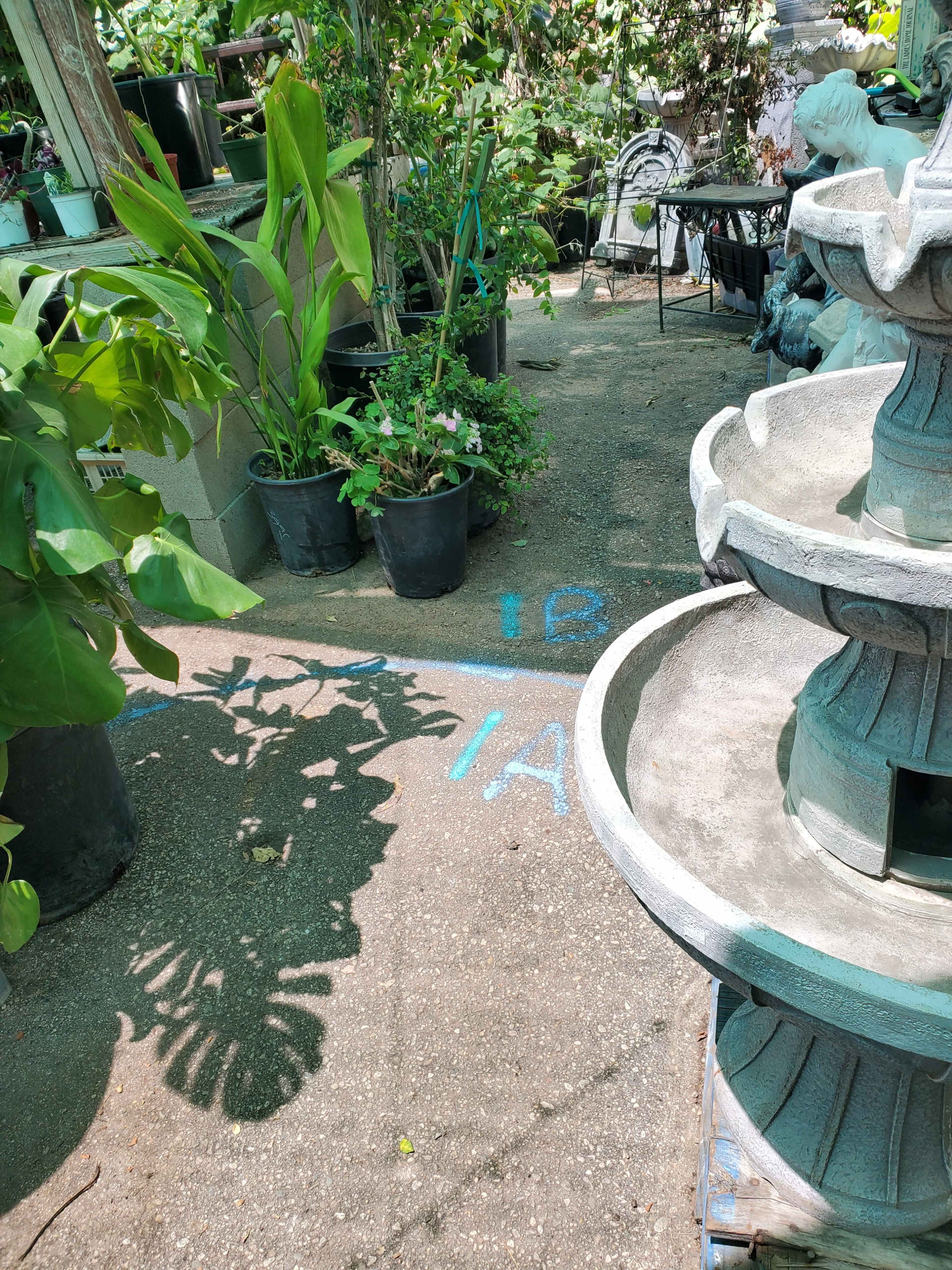 A garden path lined with potted plants and a stone fountain with shadows cast on the ground.