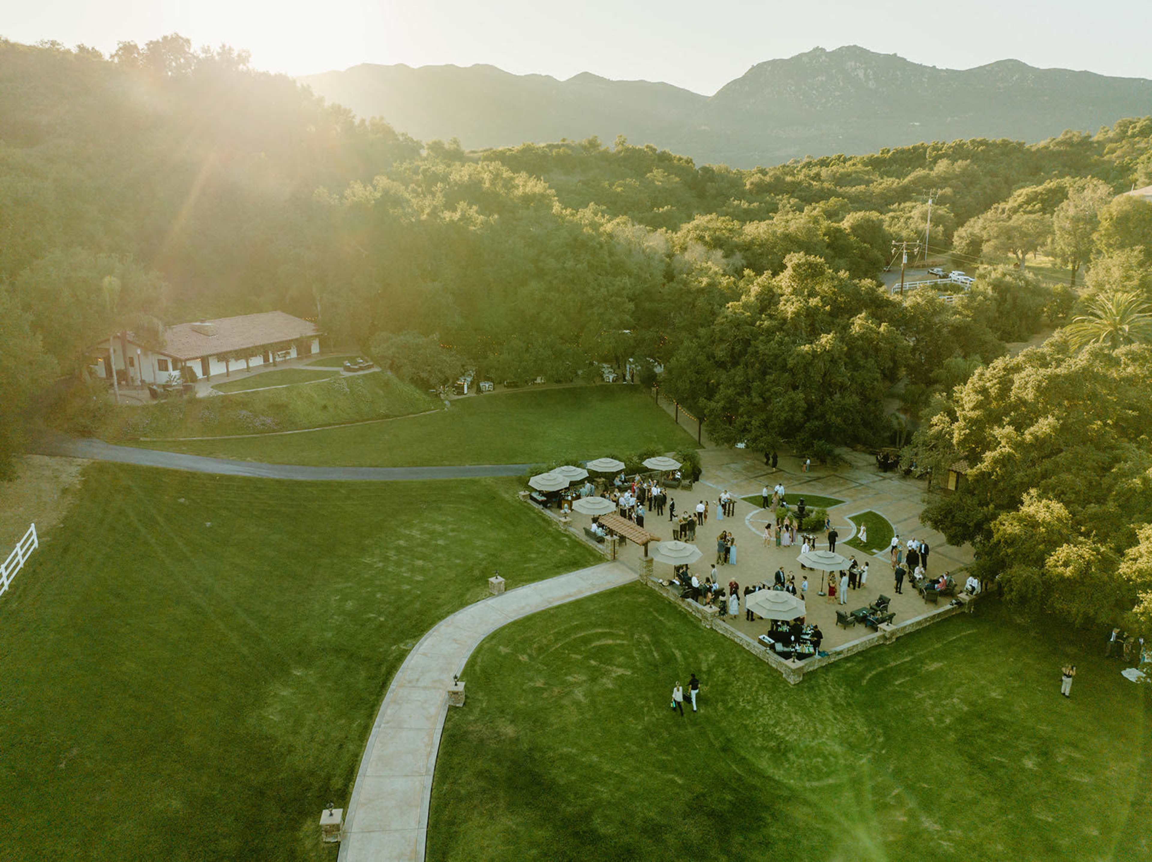 An aerial view shows a large outdoor gathering at a venue surrounded by trees and mountains in the background.