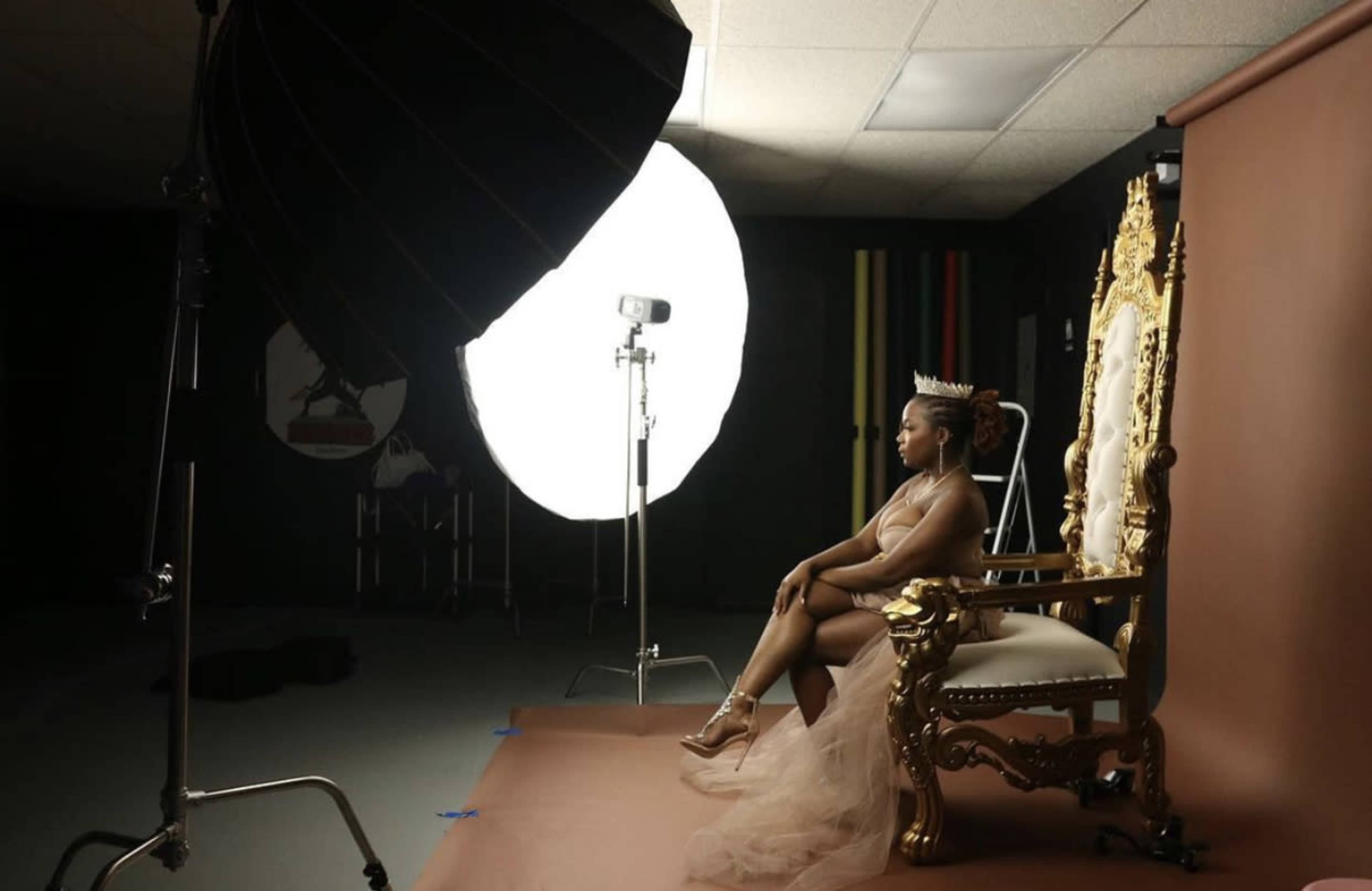A woman sits on an ornate throne in a photography studio, with a large softbox light illuminating the scene.