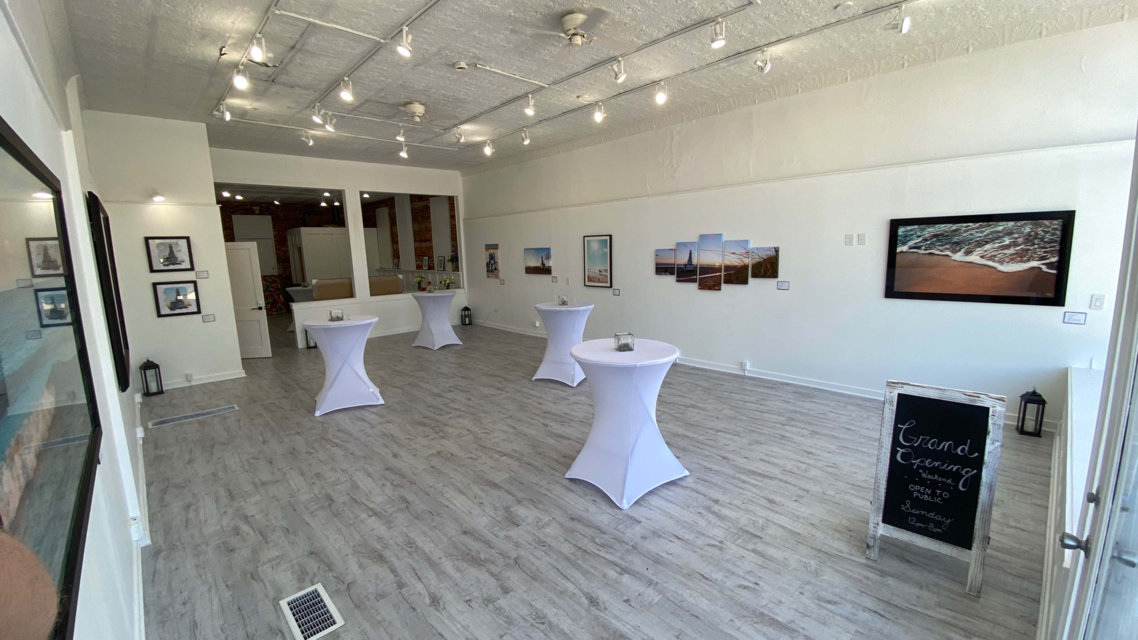 The image shows a spacious, well-lit gallery featuring several white cocktail tables, framed photographs on the walls, and a chalkboard sign promoting a grand opening event.