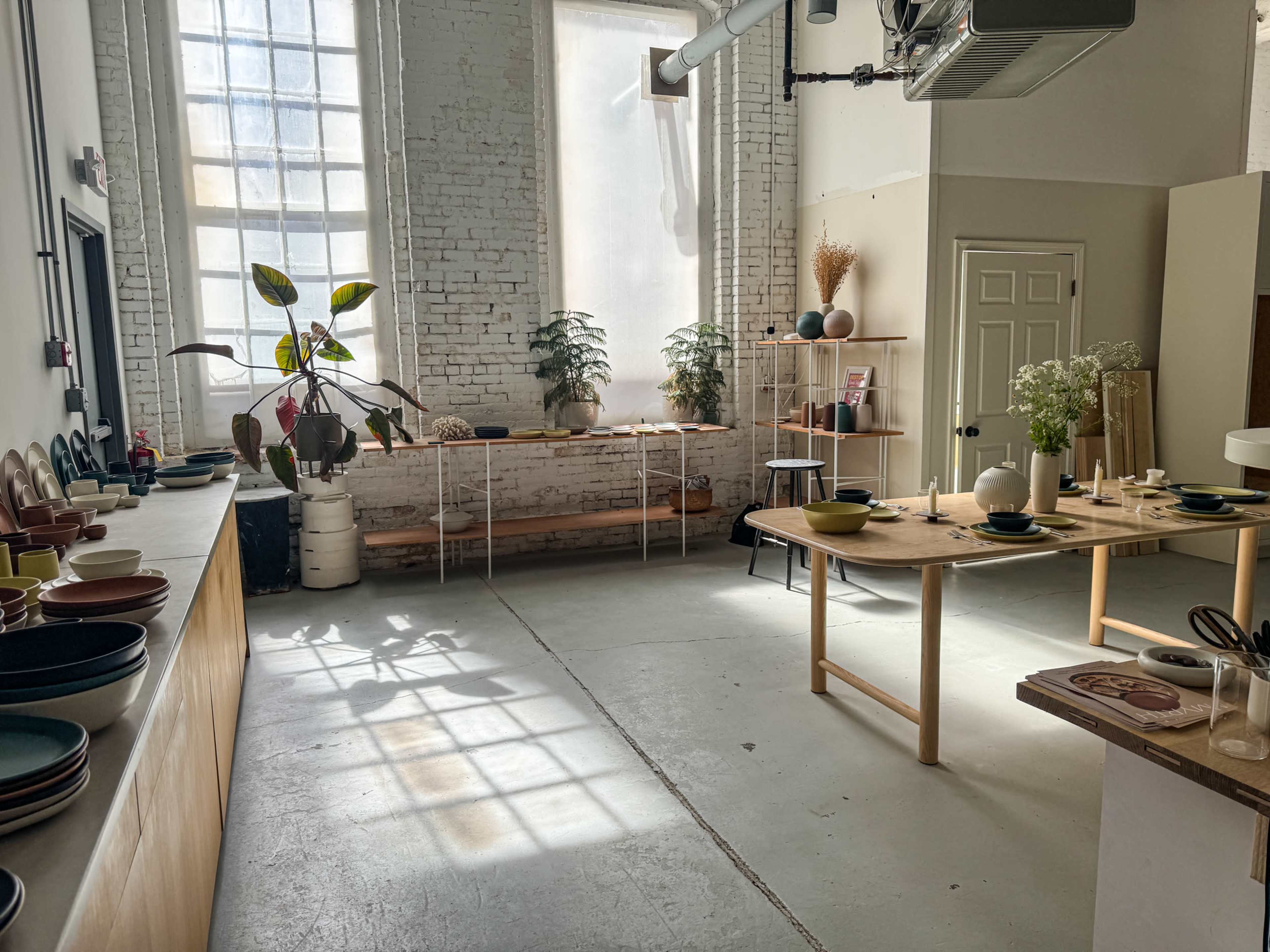 The image shows a bright and spacious interior of a showroom, featuring a table set with dishes, various pottery, plants, and large windows allowing natural light to flood the room.